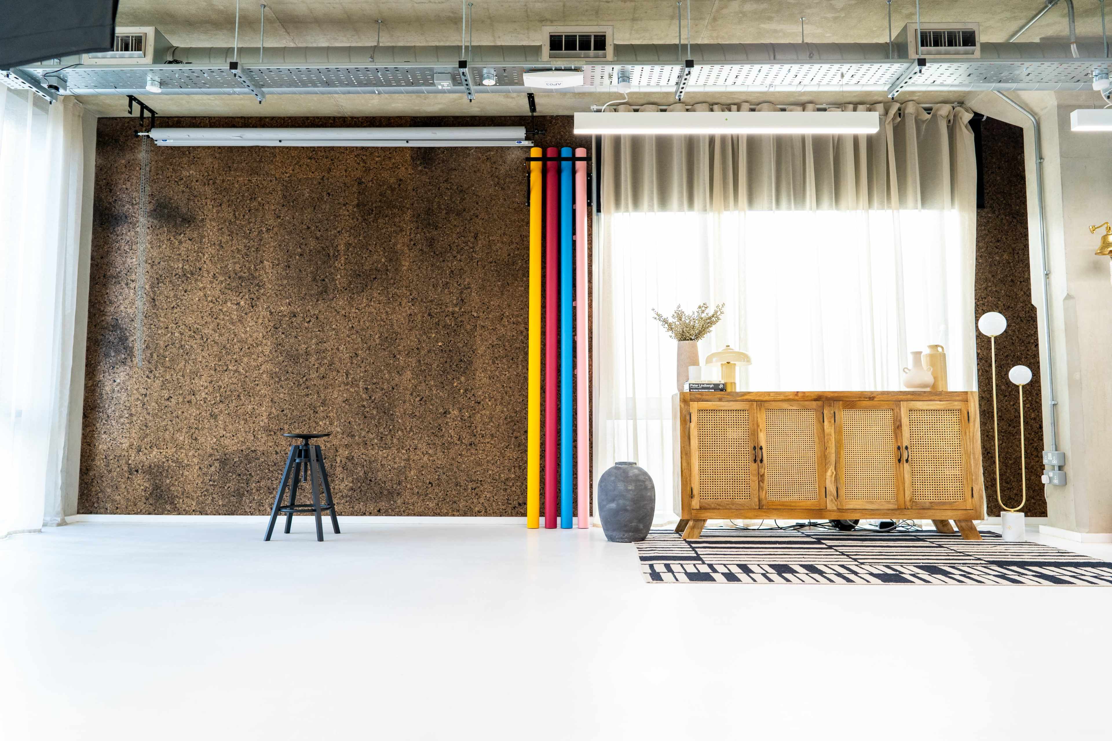 The image shows a bright, minimalist interior with a cork wall, colorful pipes, a wooden cabinet, a vase, and a patterned rug.