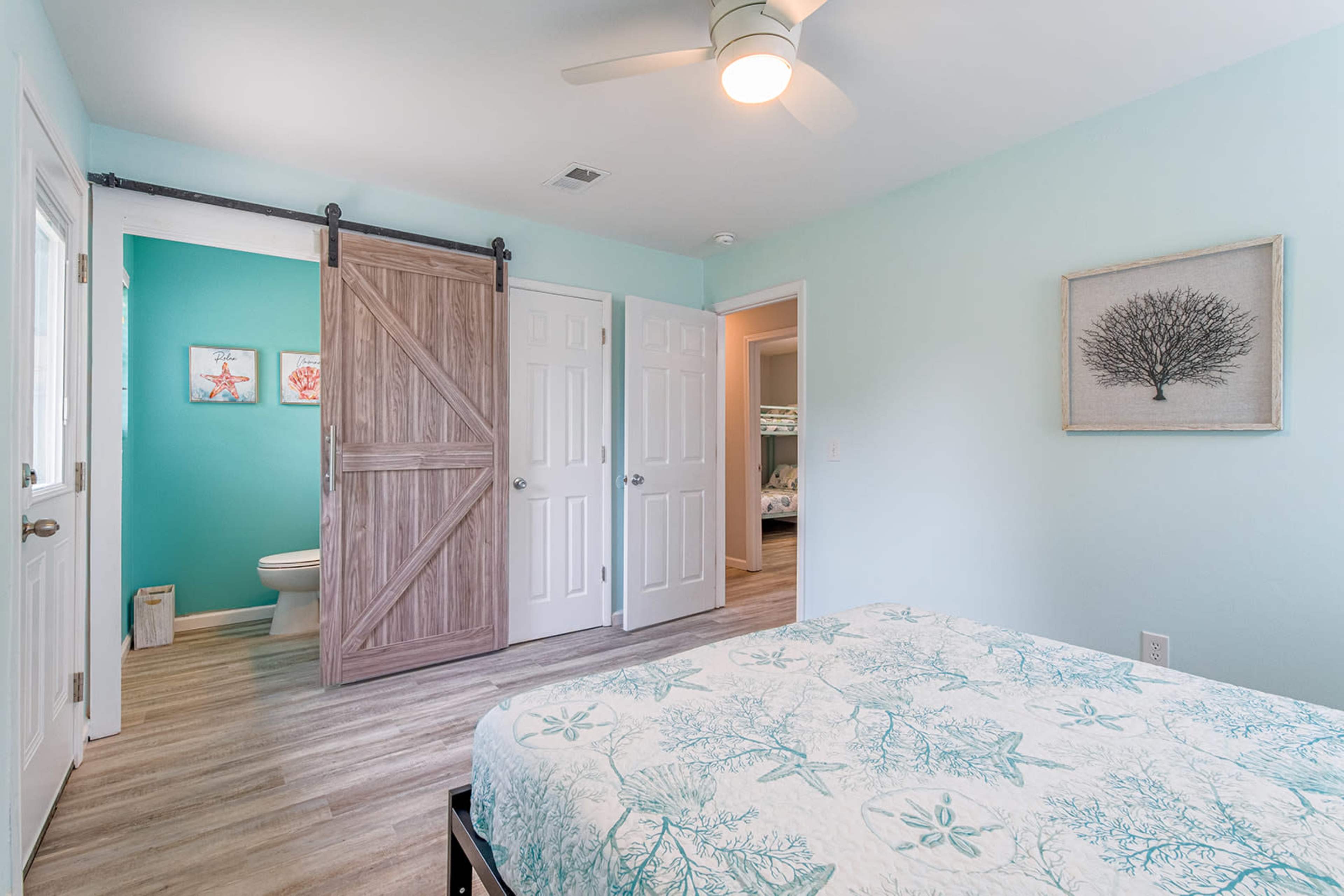 A light blue bedroom with a bed covered in a patterned quilt, leading to a bathroom visible through an open sliding barn door.
