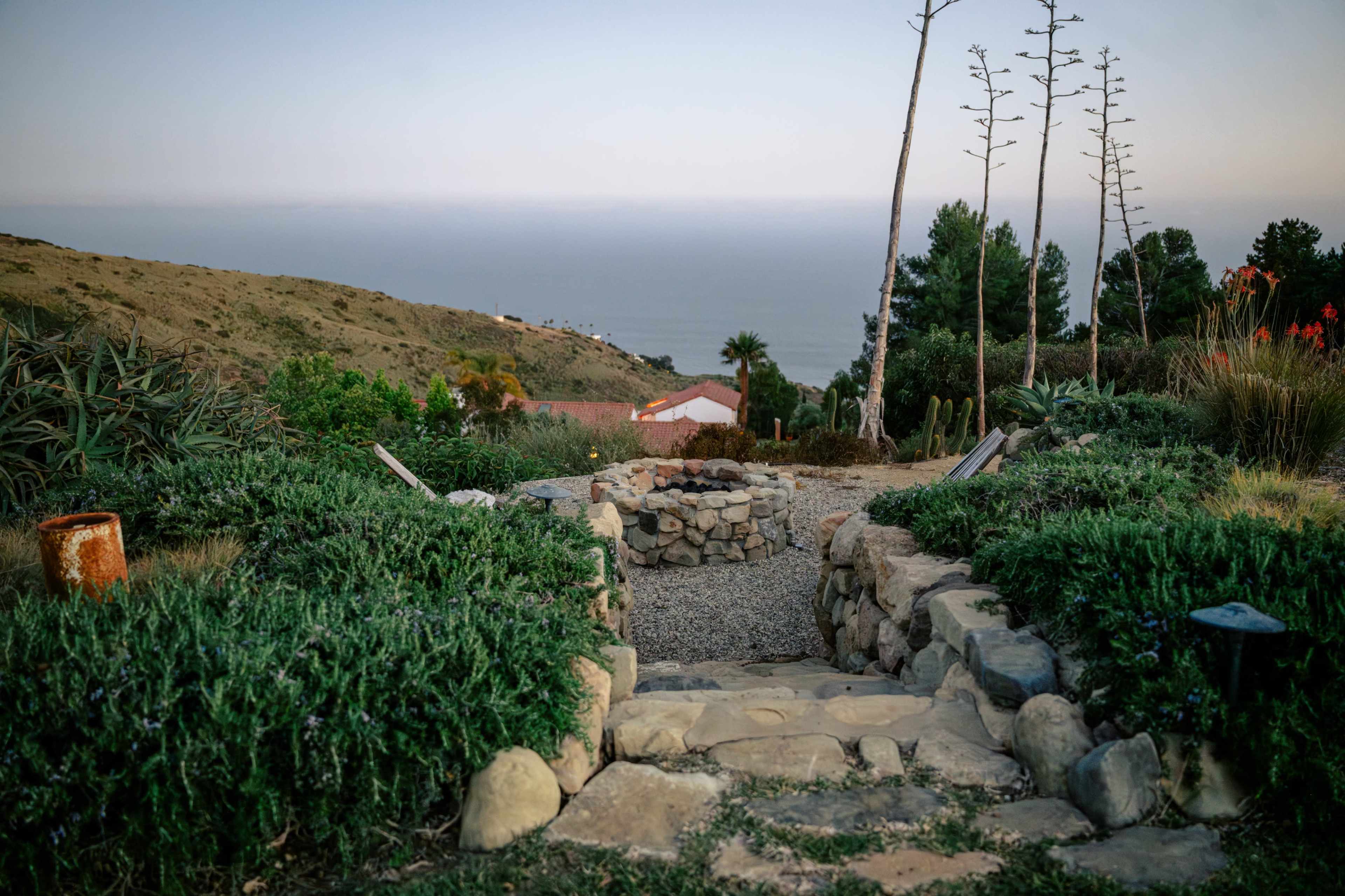 A stone pathway leads down to a circular fire pit surrounded by lush greenery, with a view of the ocean in the distance.