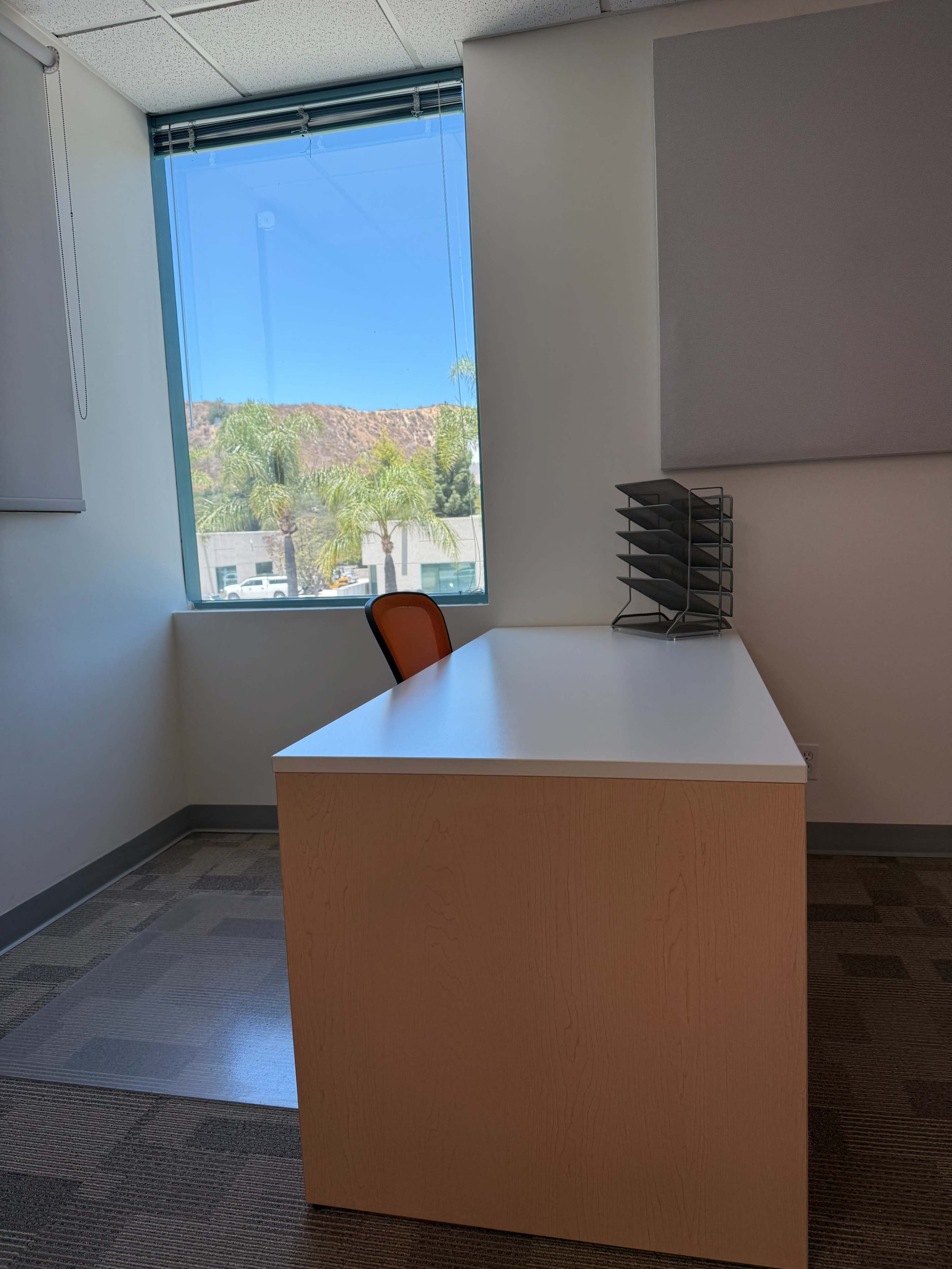 The image shows a minimalist office space with a white desk, an orange chair, and a file organizer near a window that overlooks a view of palm trees and a mountain.