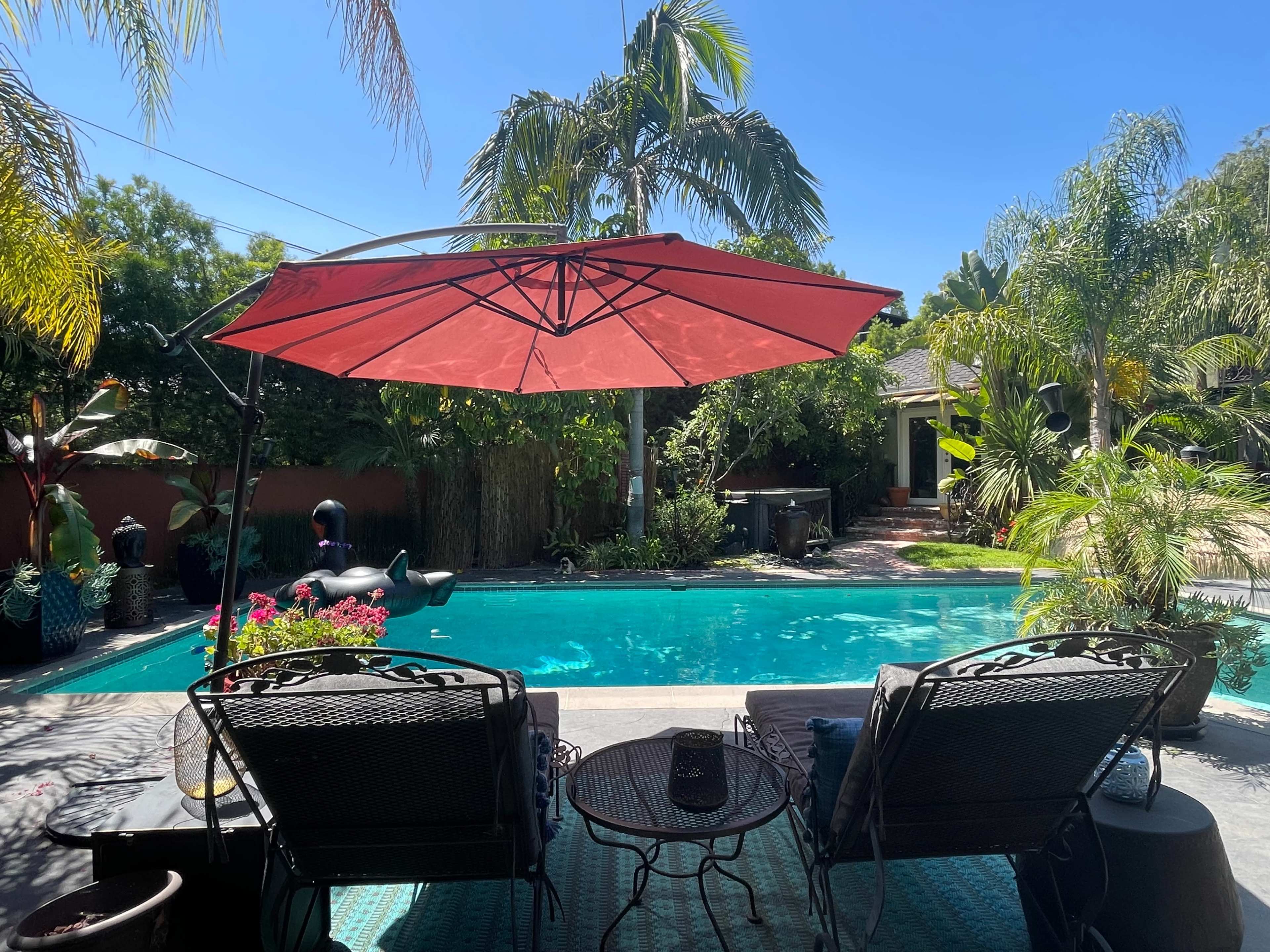A shaded outdoor seating area overlooks a swimming pool surrounded by tropical plants and trees.