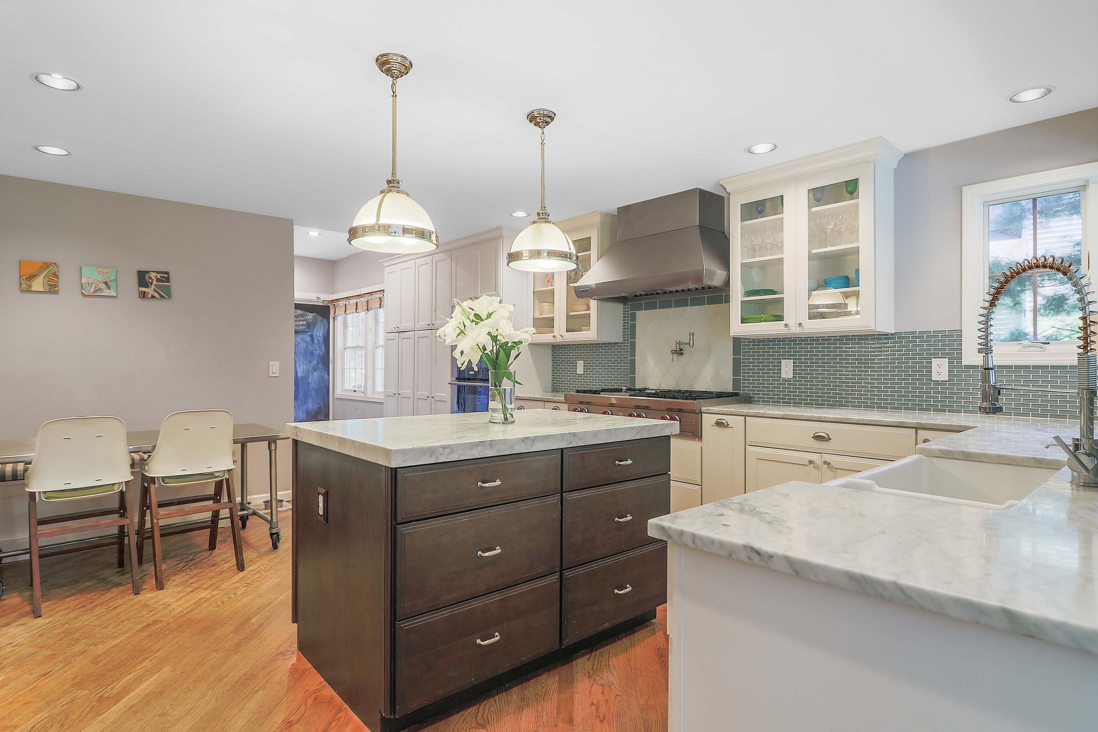 The image shows a modern kitchen with a central island, wooden floors, stainless steel appliances, and light fixtures hanging above.