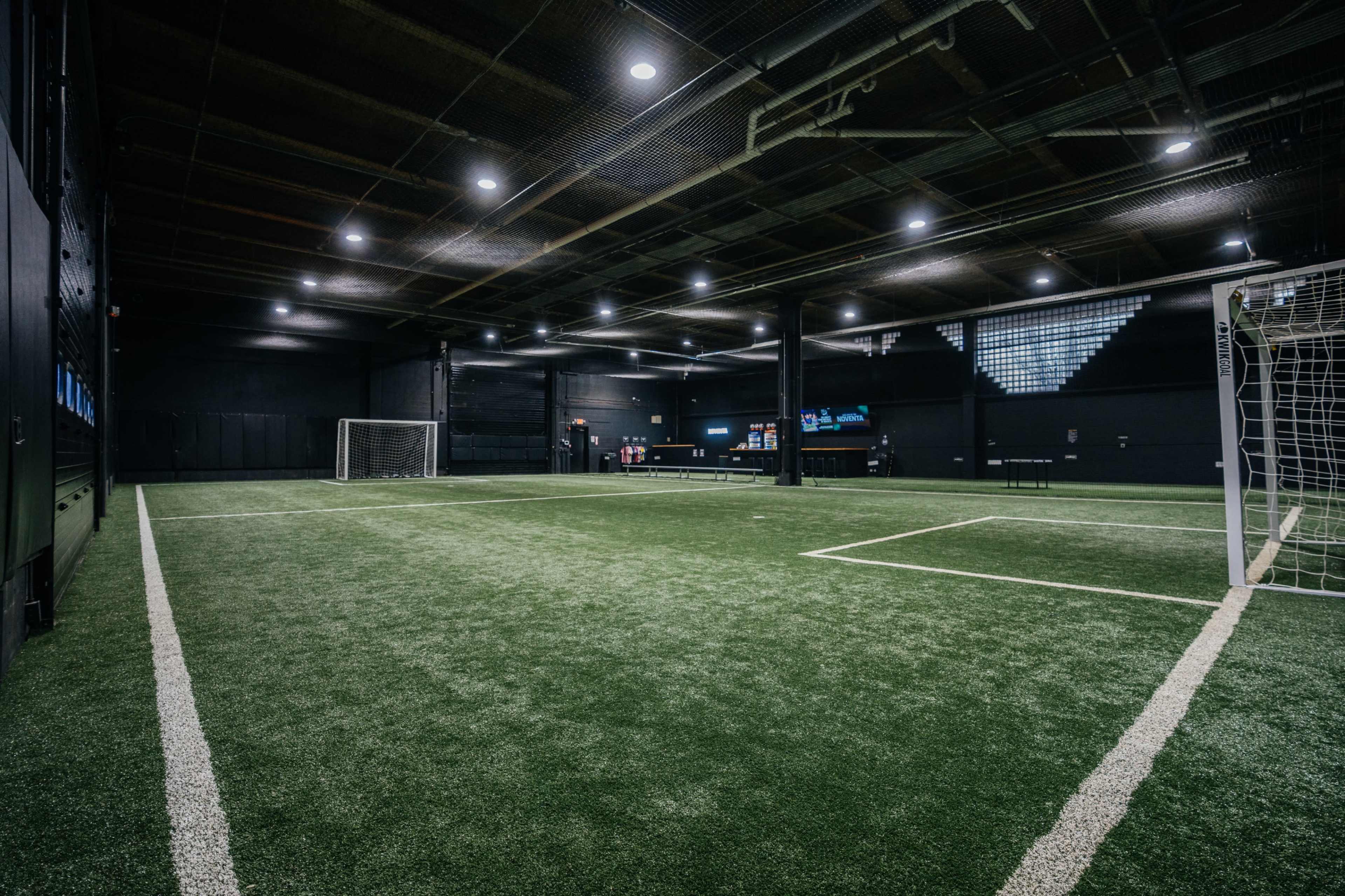 The image shows an indoor soccer field with artificial turf and goals at either end, illuminated by overhead lights.