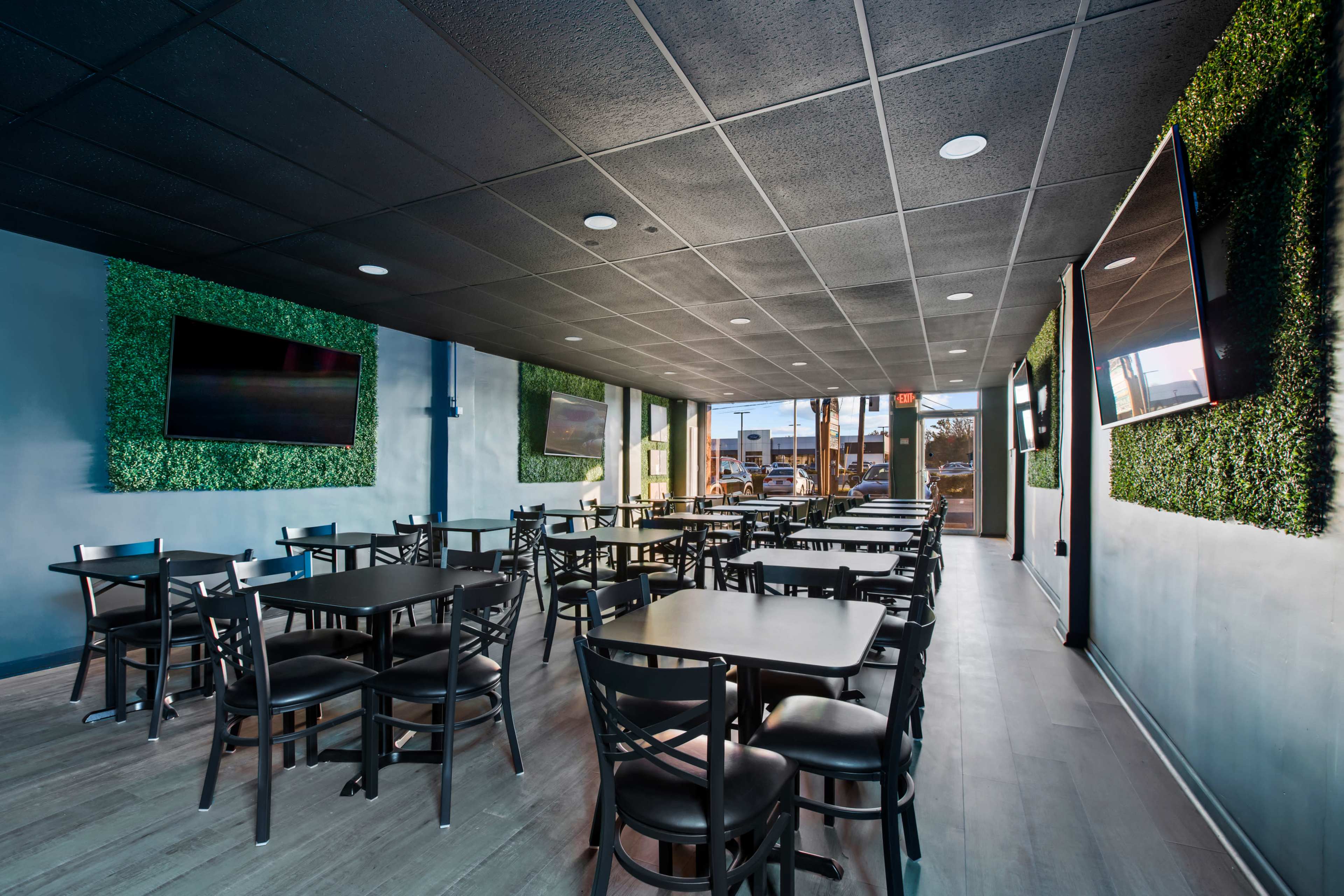 A modern restaurant interior with black tables and chairs, greenery on the walls, and large windows allowing natural light to enter.