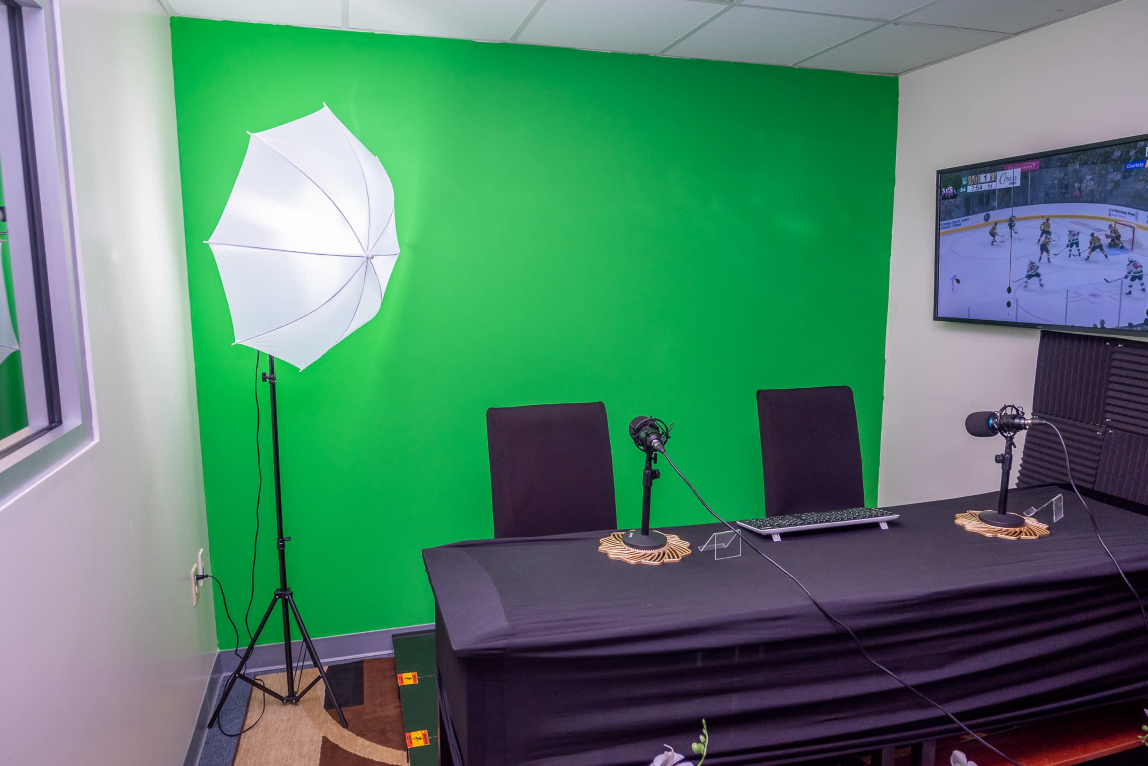 A small studio with a green screen wall, two chairs at a table, microphones on stands, and a monitor displaying a sports game.
