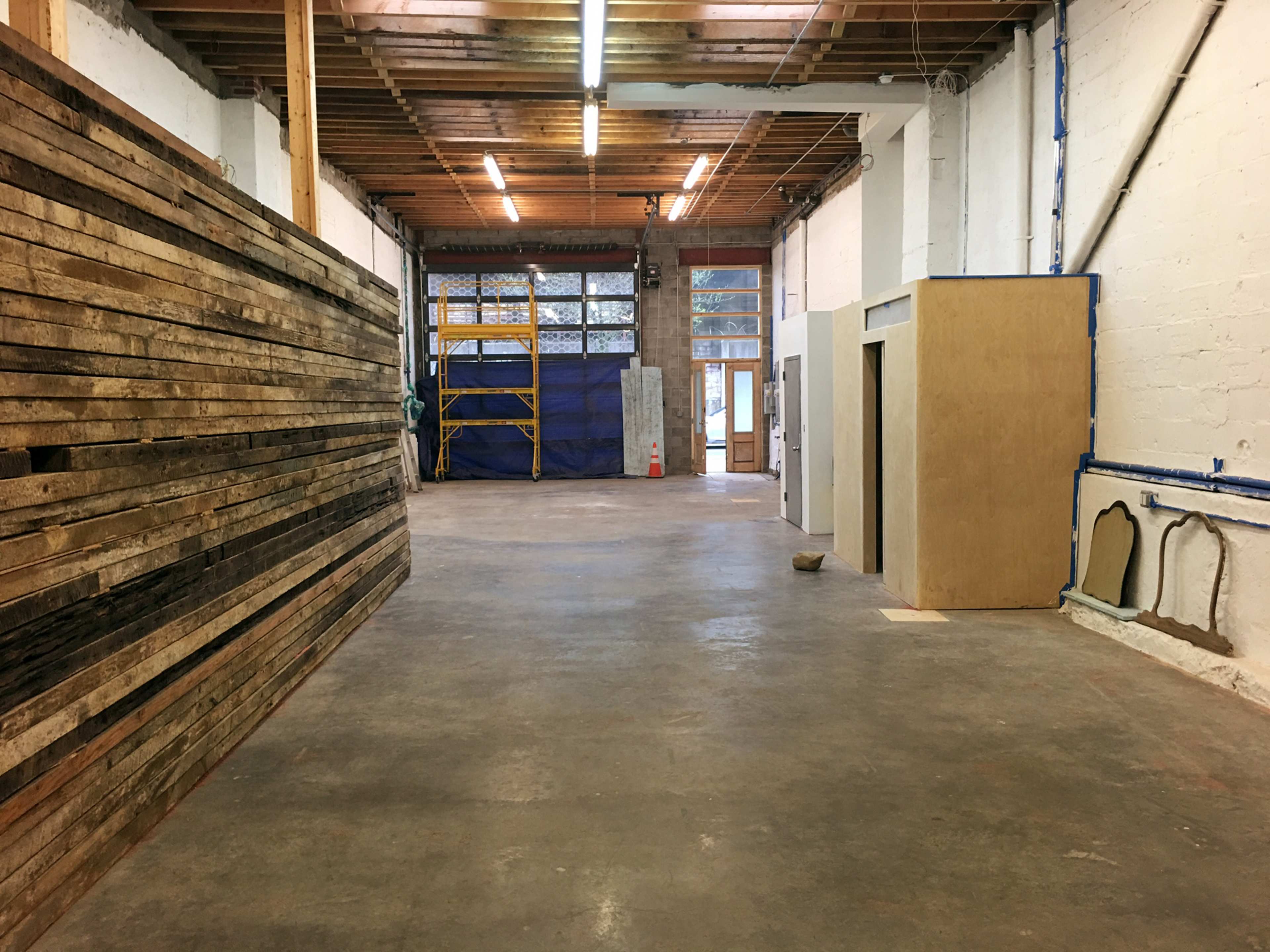 The image shows a spacious interior of a workshop or construction area with wooden walls on one side, an unfinished wall space, and scaffolding in the background.