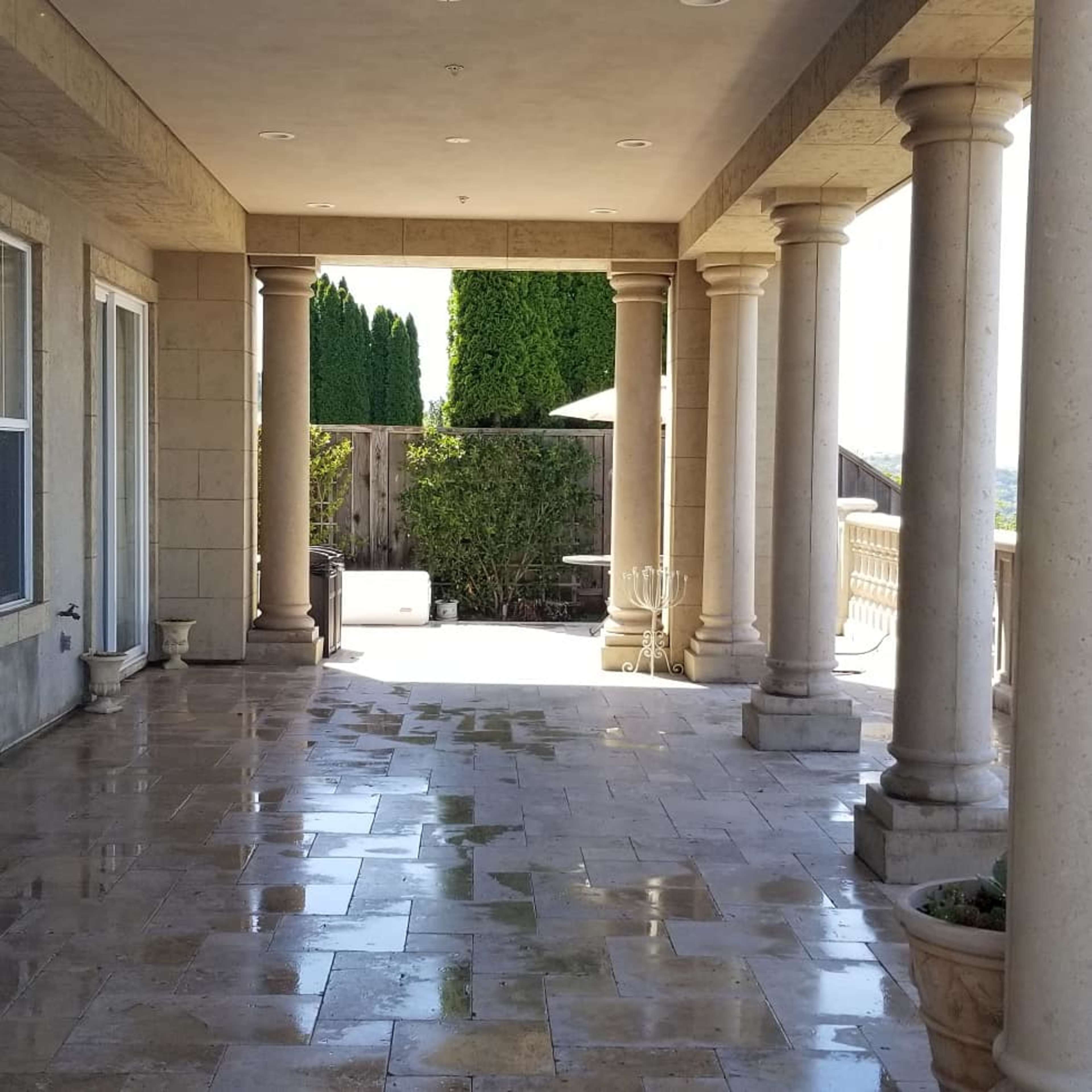A covered patio features stone columns and a tiled floor, with greenery visible in the background.