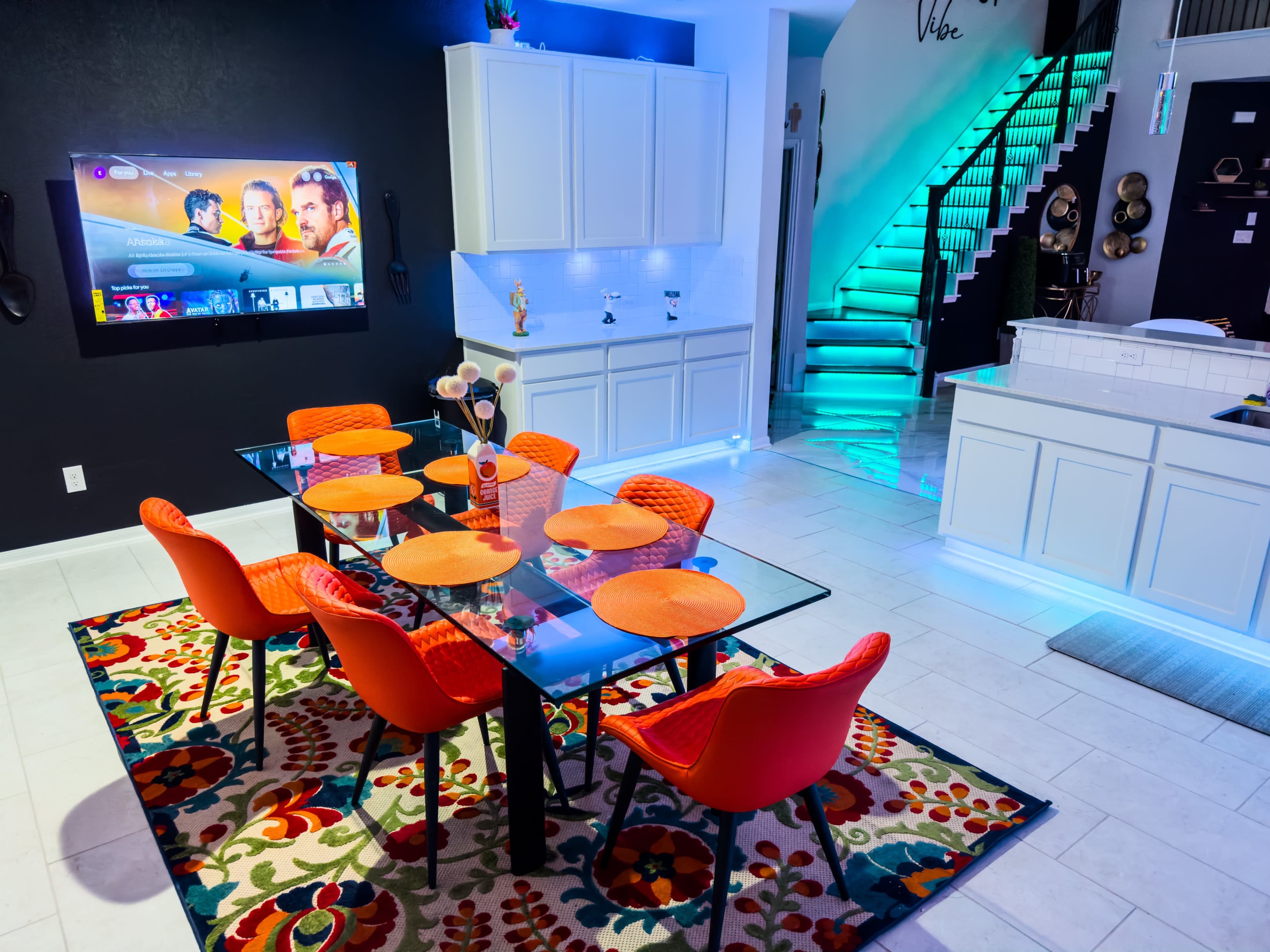 A modern dining area with a glass table surrounded by orange chairs, a rug with a floral pattern, and a kitchen in the background featuring white cabinetry and ambient lighting.