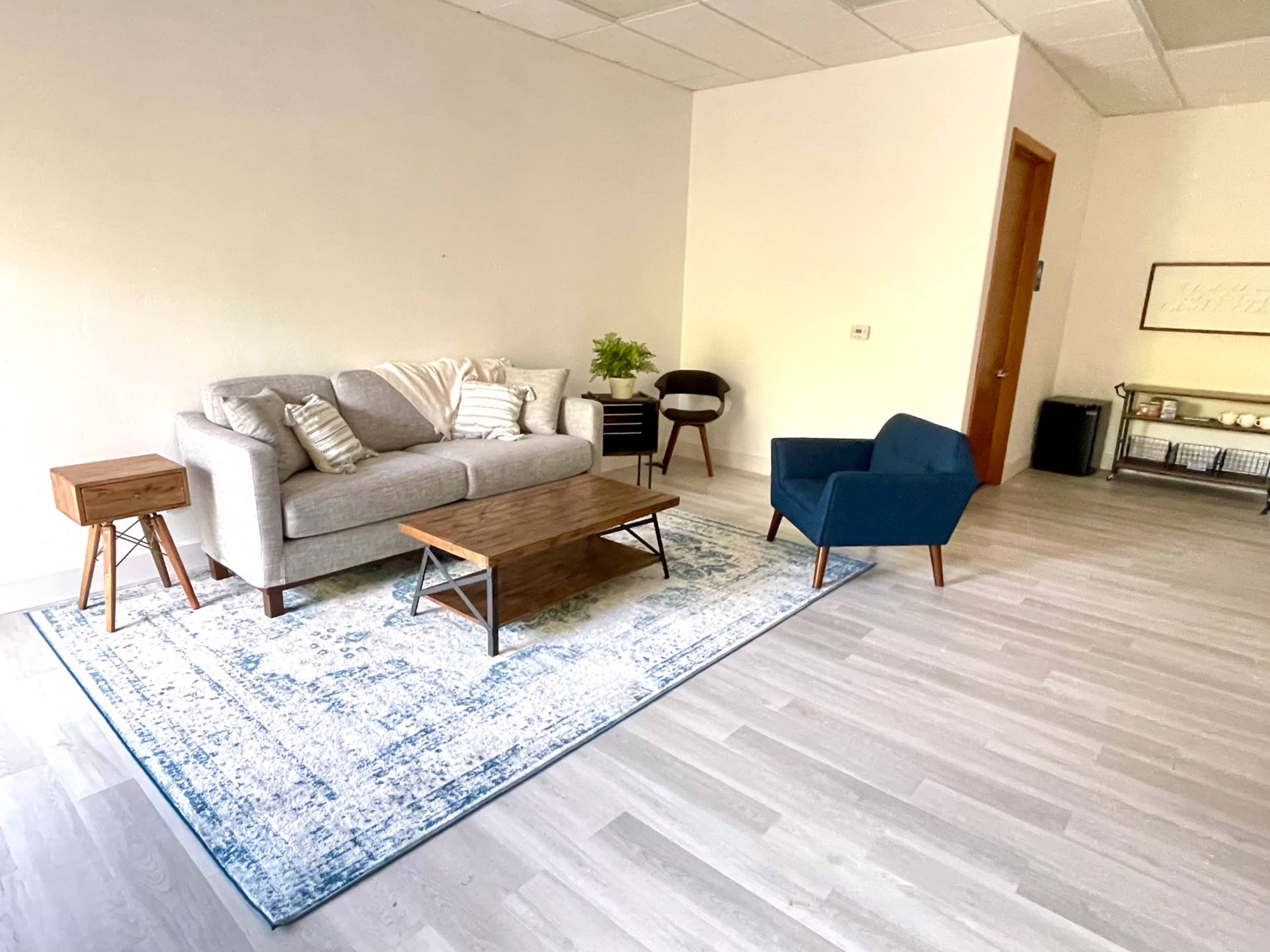 A living room features a gray sofa, a blue accent chair, a wooden coffee table, and a large area rug on a light-colored floor.