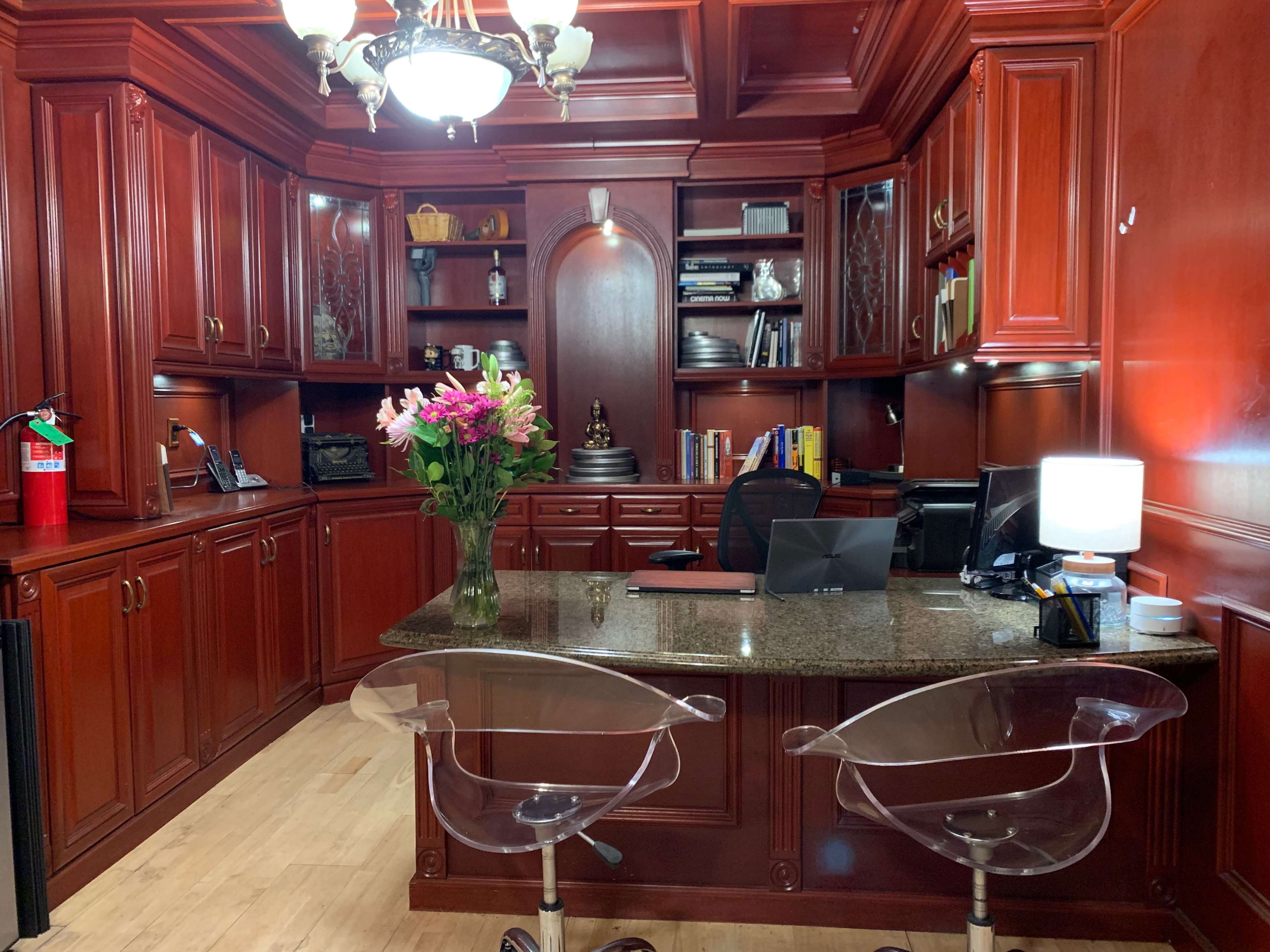 A wood-paneled office with a granite countertop, clear acrylic chairs, and shelves filled with books and decorative items, illuminated by a chandelier.