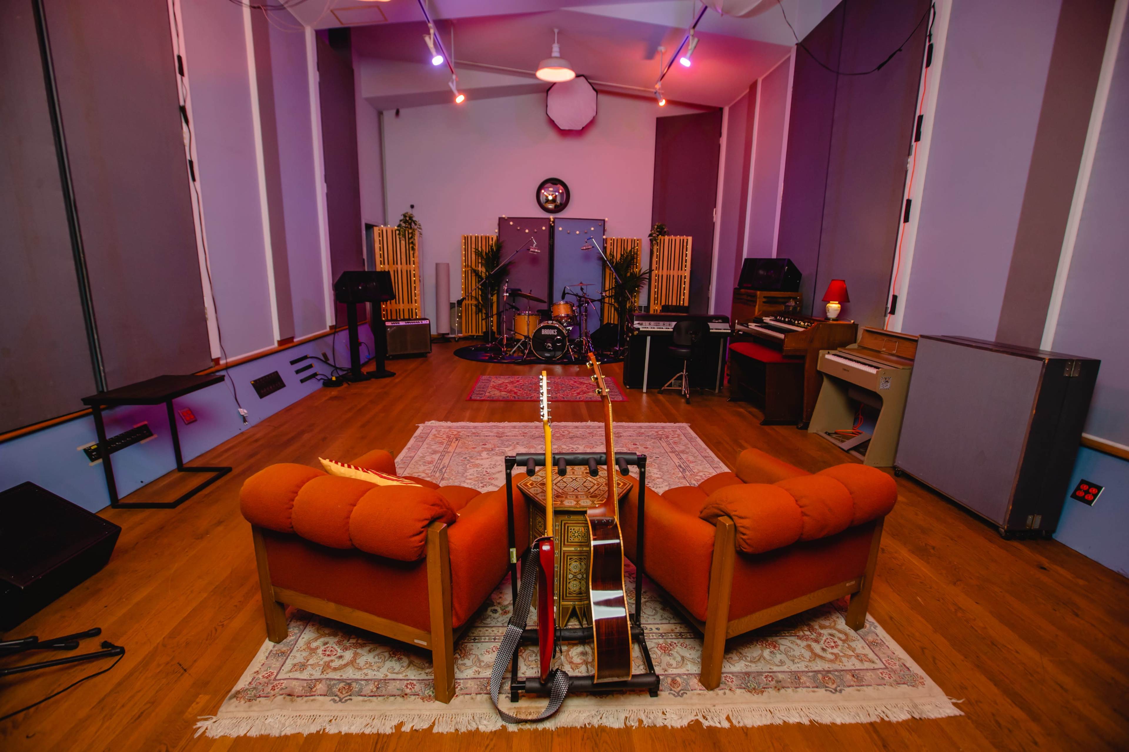 A cozy music studio features two red armchairs facing a guitar and rug, with a drum set and amplifiers in the background.
