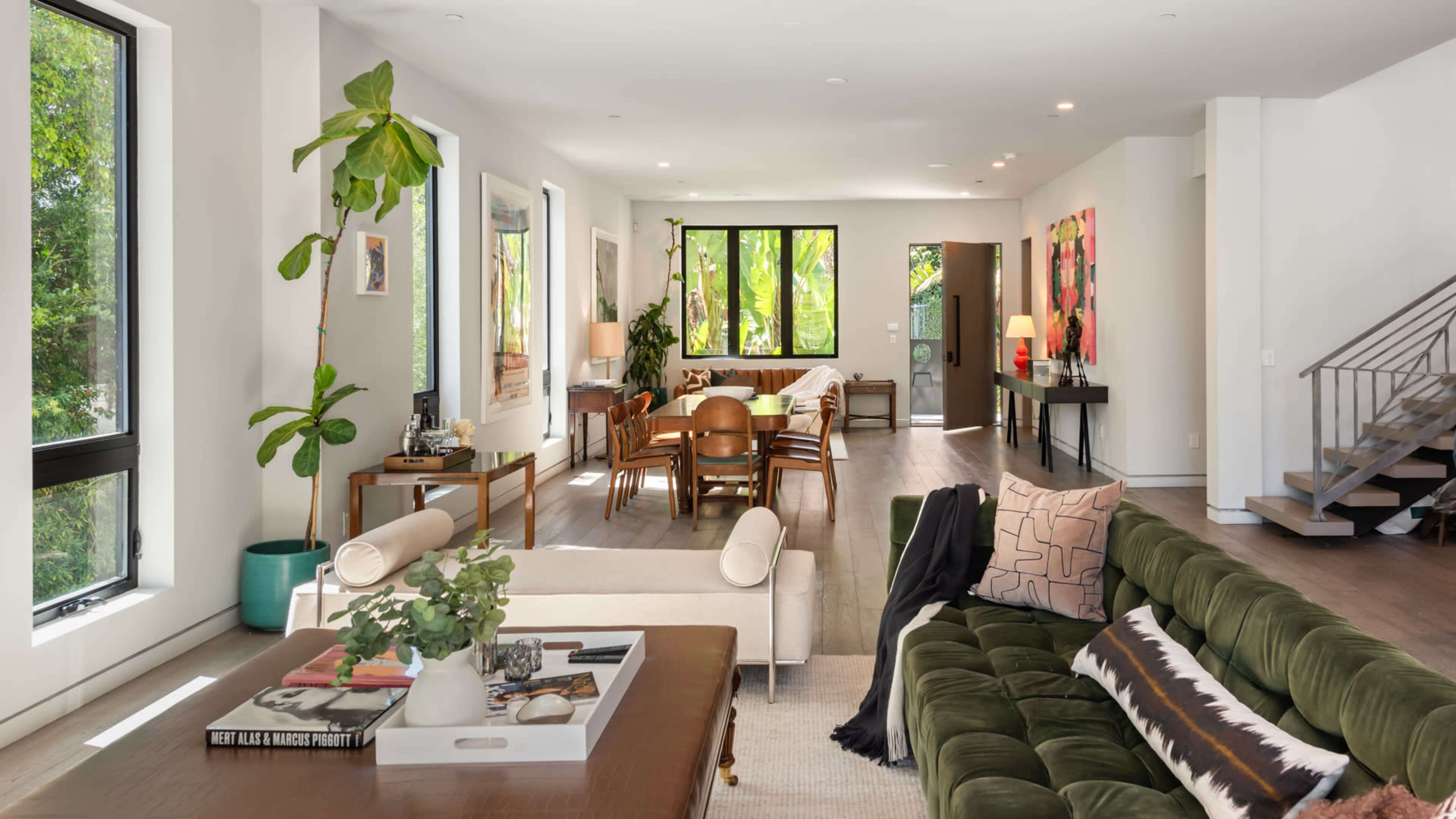 A spacious living area features a dark green sectional sofa, a wooden coffee table with plants, and a dining table surrounded by chairs, all illuminated by natural light from large windows.