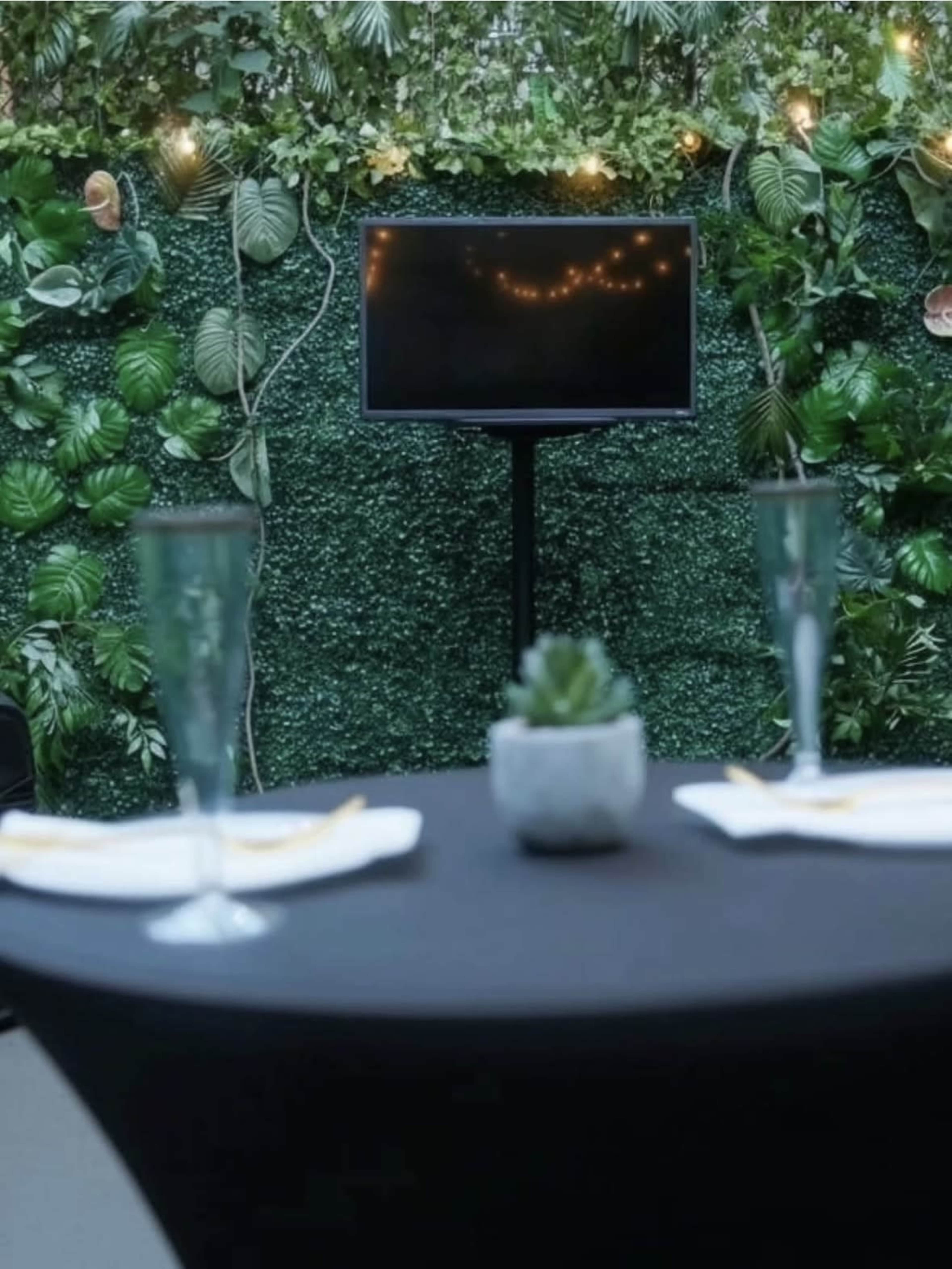 A table set for a meal is positioned in front of a green, plant-covered backdrop with a screen displaying soft lights.
