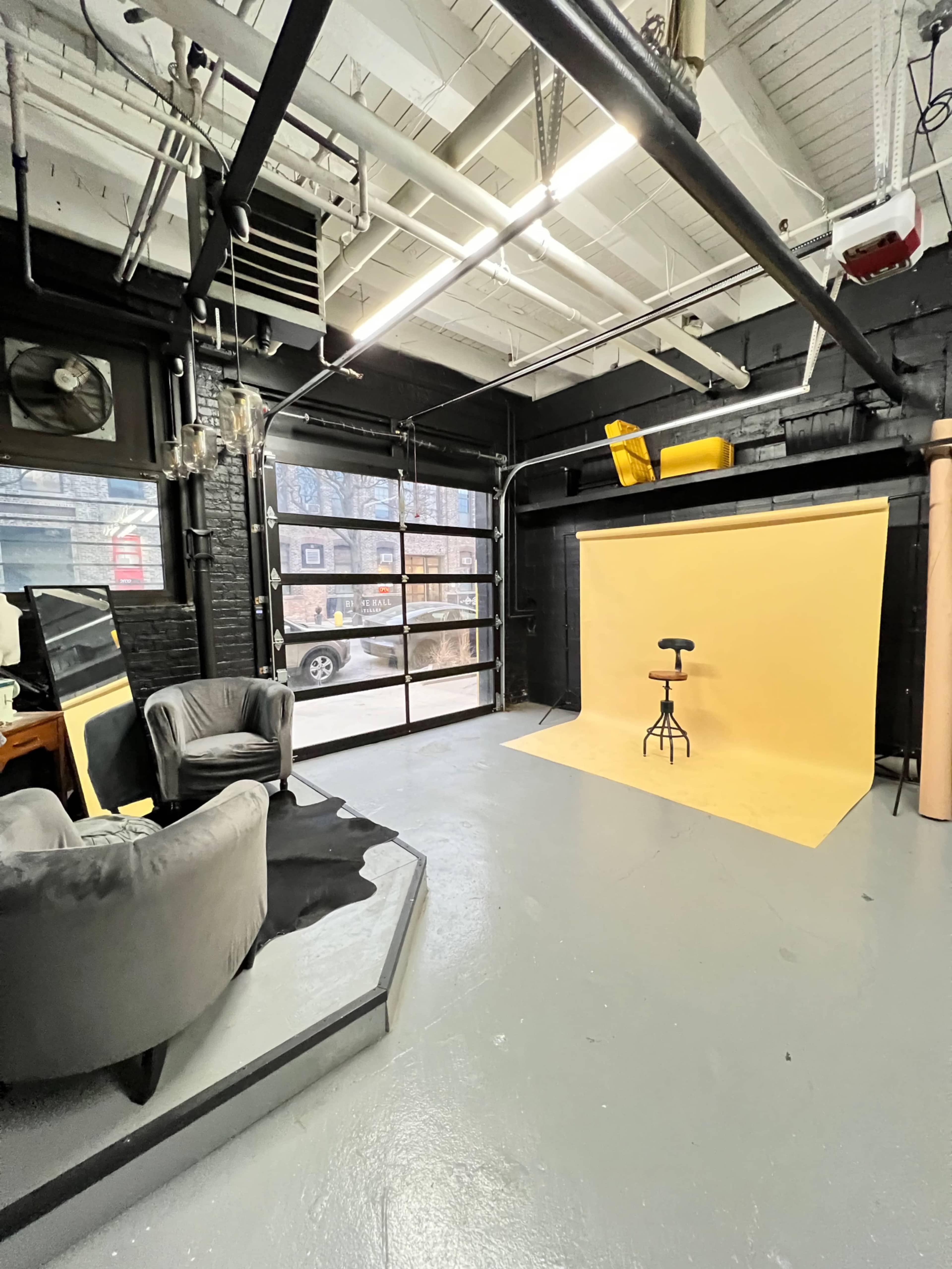 A spacious, modern studio with a yellow backdrop set up for photography, featuring a stool on the backdrop and seating areas in the foreground.