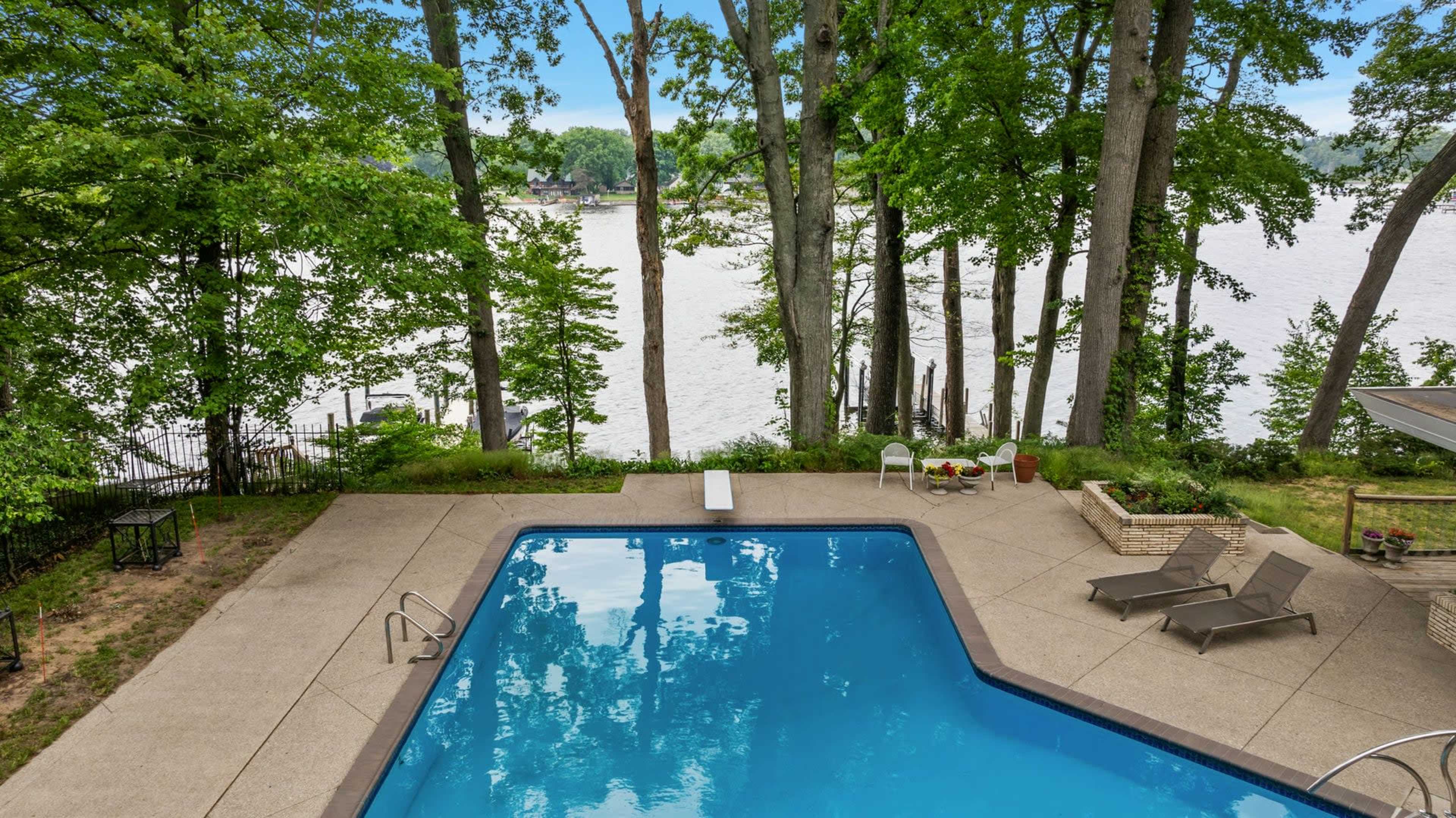 A swimming pool is situated beside a lake, surrounded by tall trees and outdoor furniture.