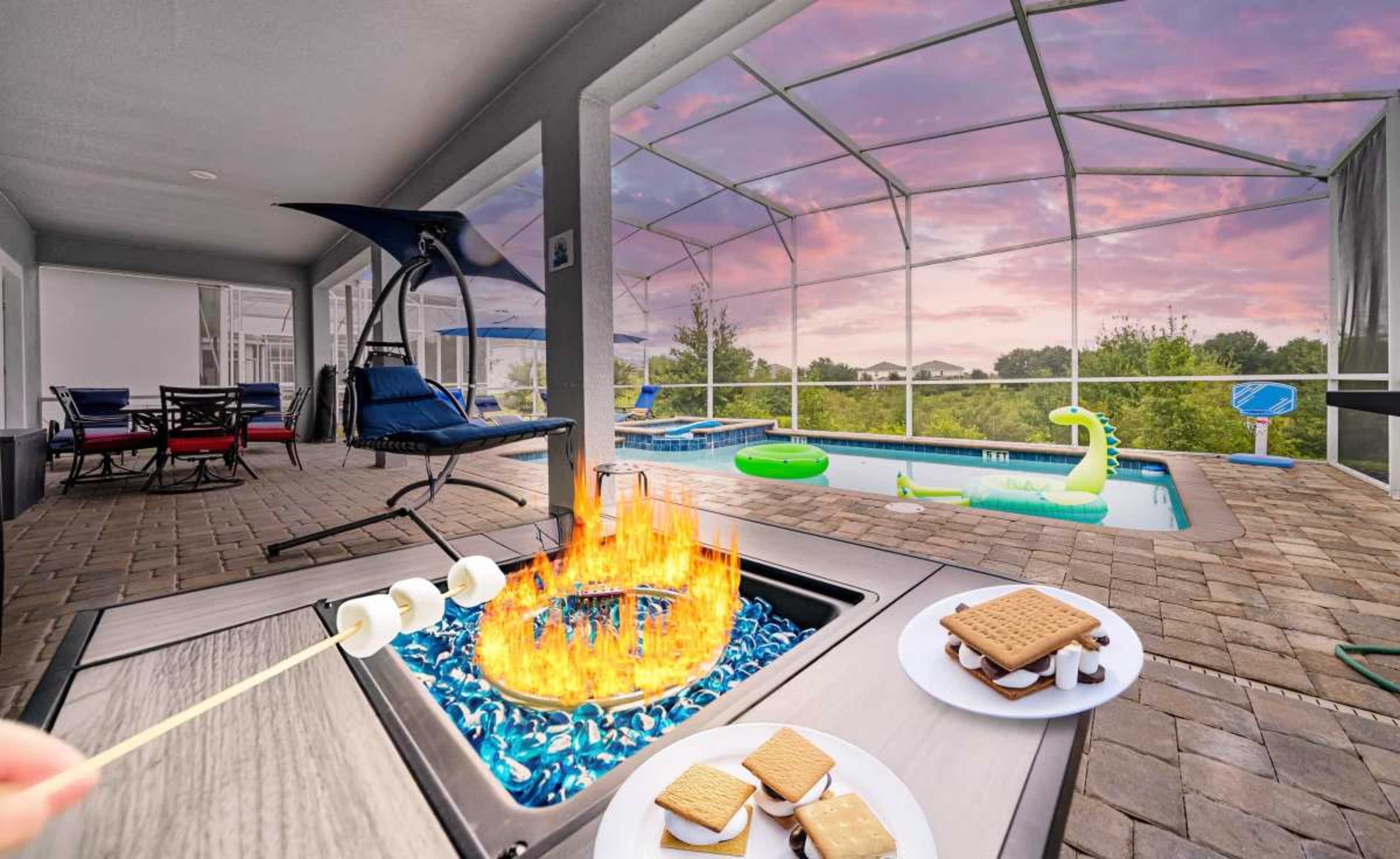 A fire pit with flames is surrounded by s'mores ingredients, with a view of a screened-in patio and a swimming pool in the background.