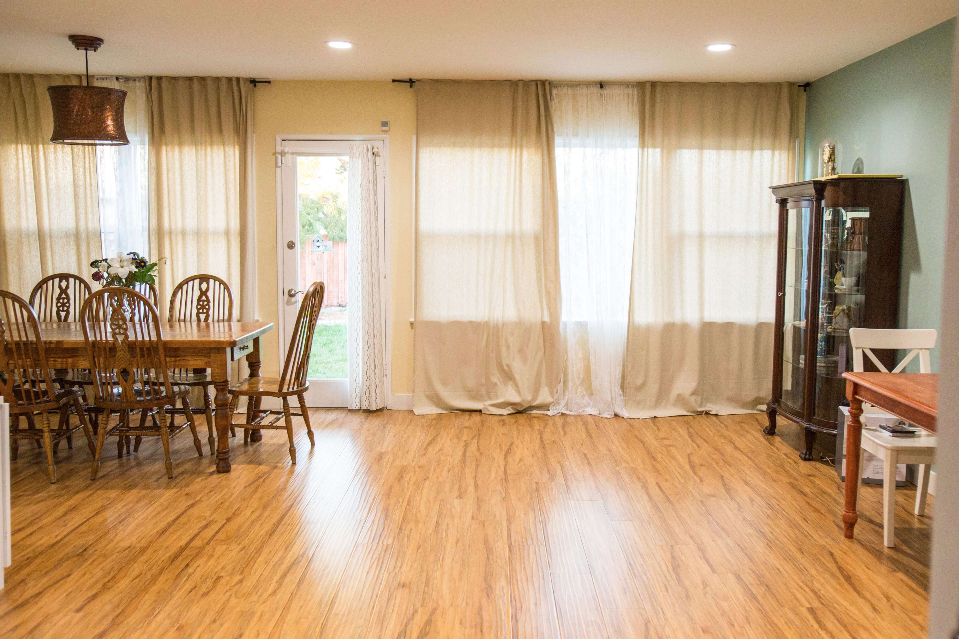 A dining area with a wooden table and chairs, a china cabinet, and large windows covered with sheer curtains.