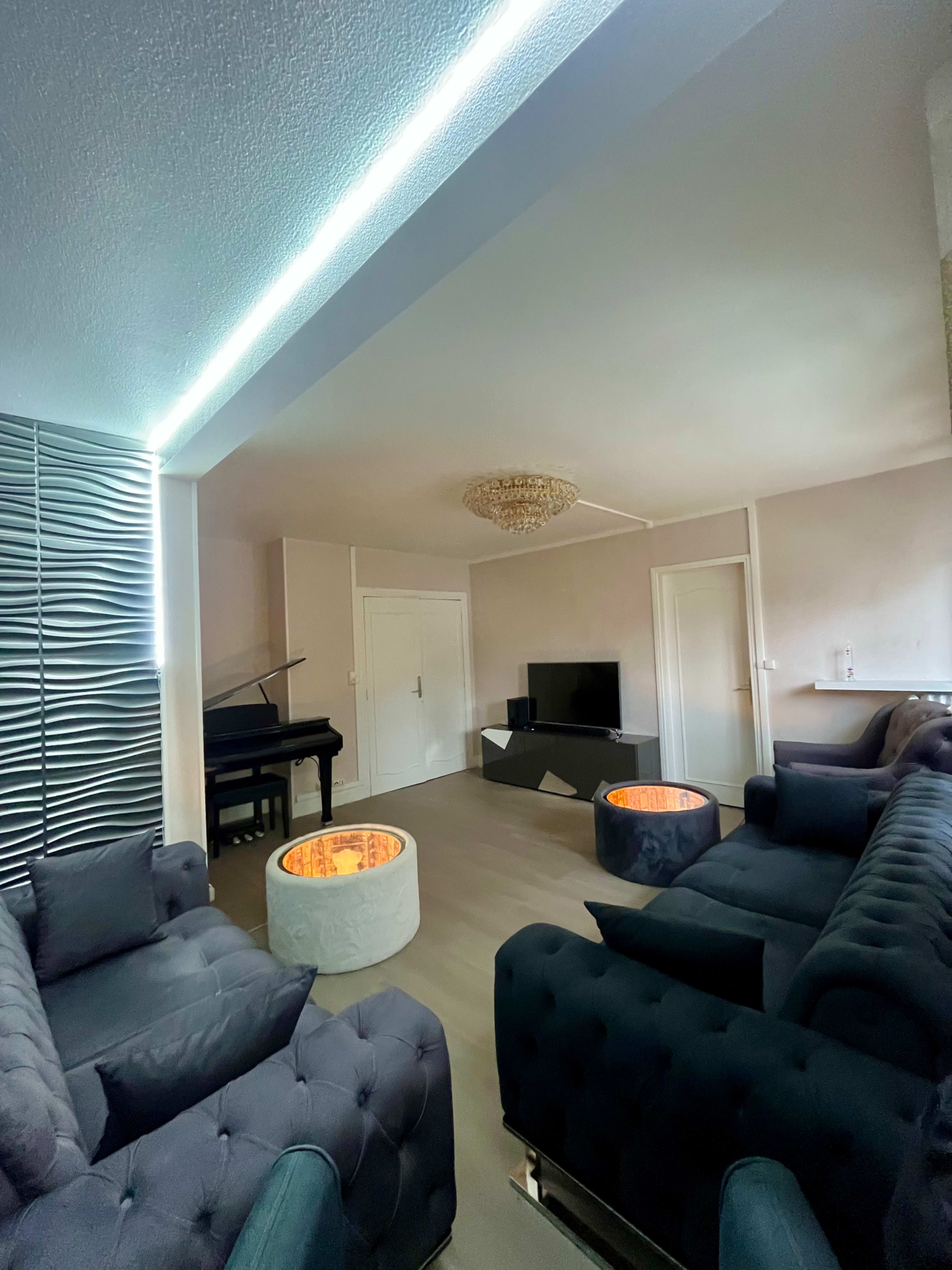 The image shows a modern living room featuring a piano, a television, and two circular coffee tables with glowing inserts, surrounded by plush seating.