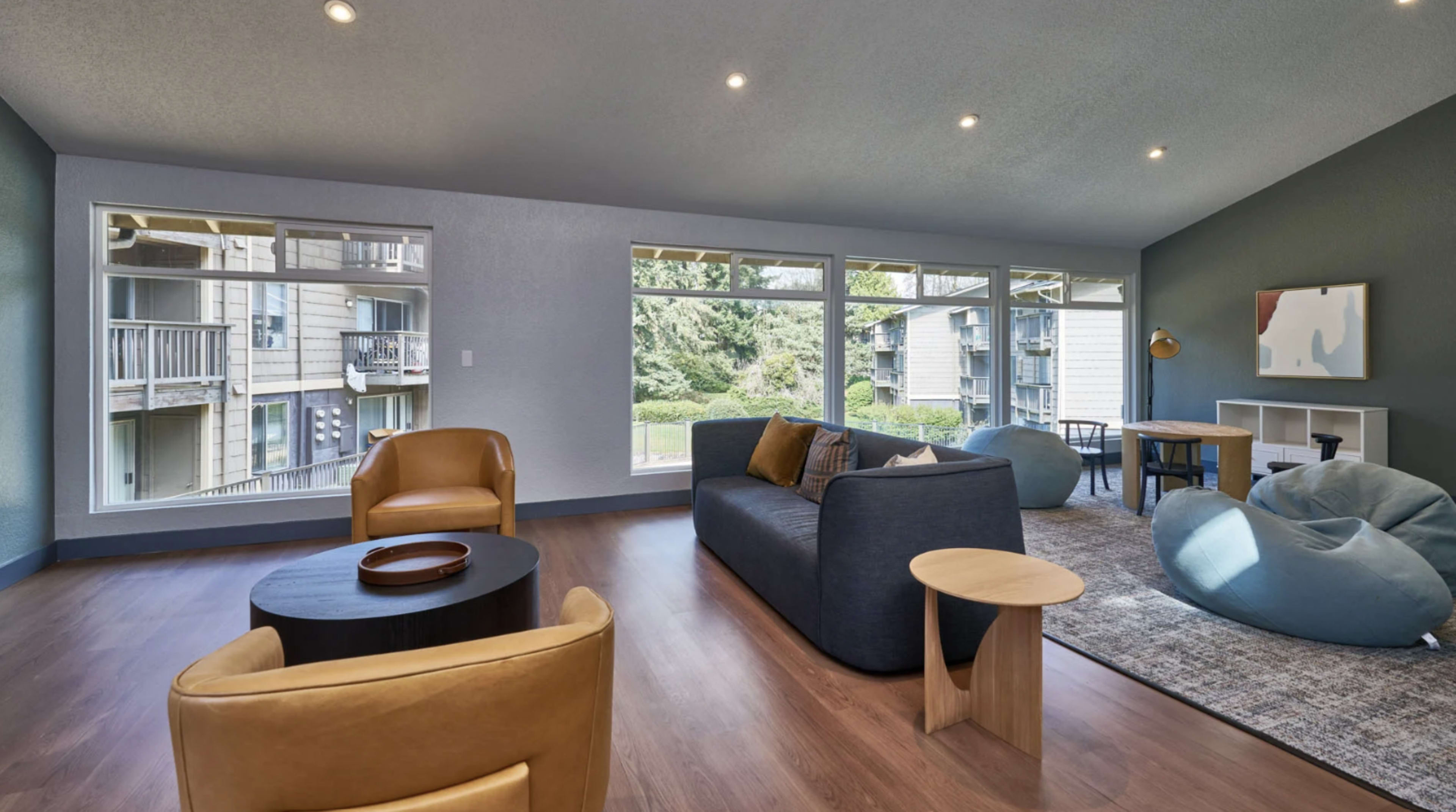 The living area features a modern design with a dark sofa, beige armchairs, a round table, and large windows overlooking greenery.