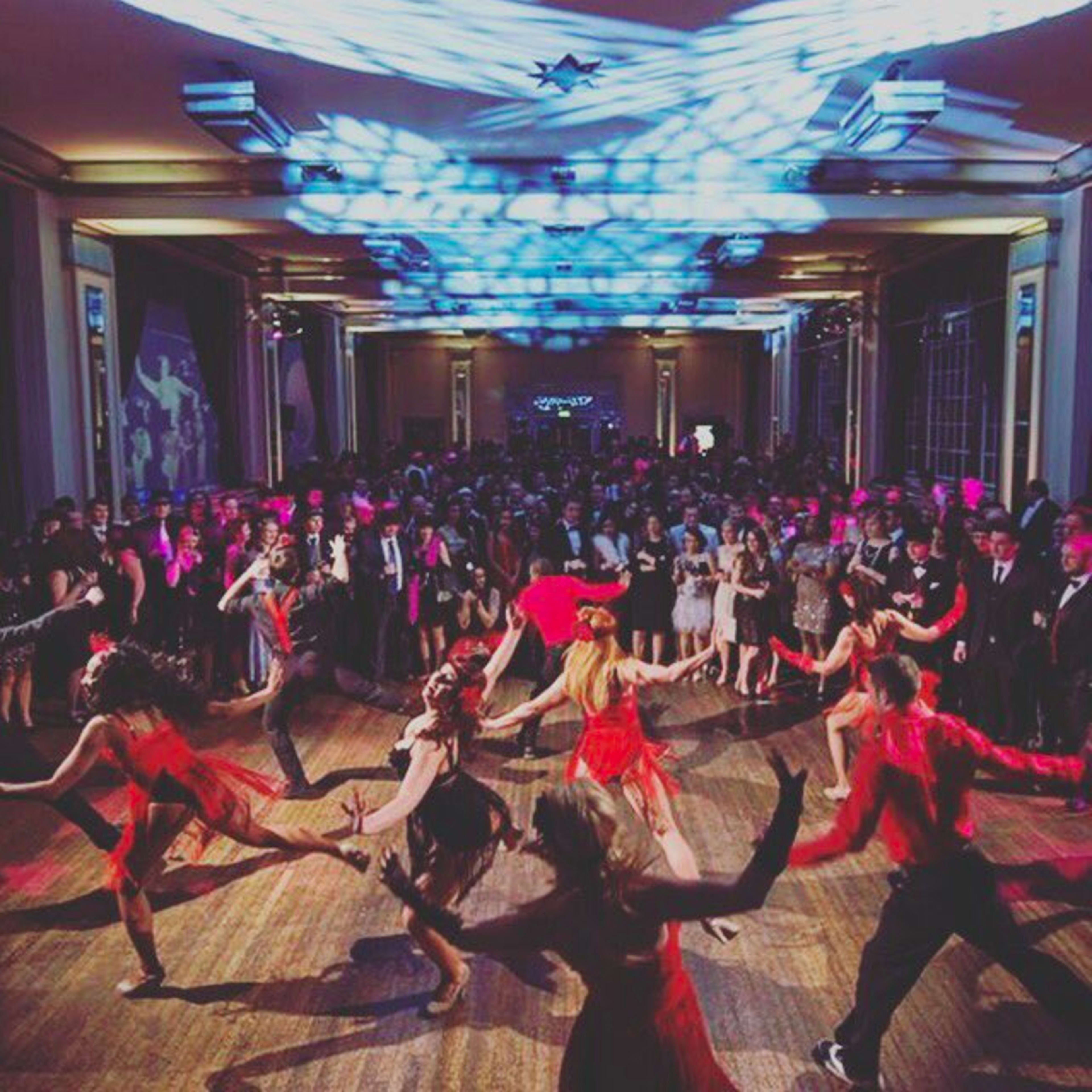 A vibrant dance performance takes place in a crowded ballroom, with dancers in colorful outfits showcasing their moves under dramatic lighting.