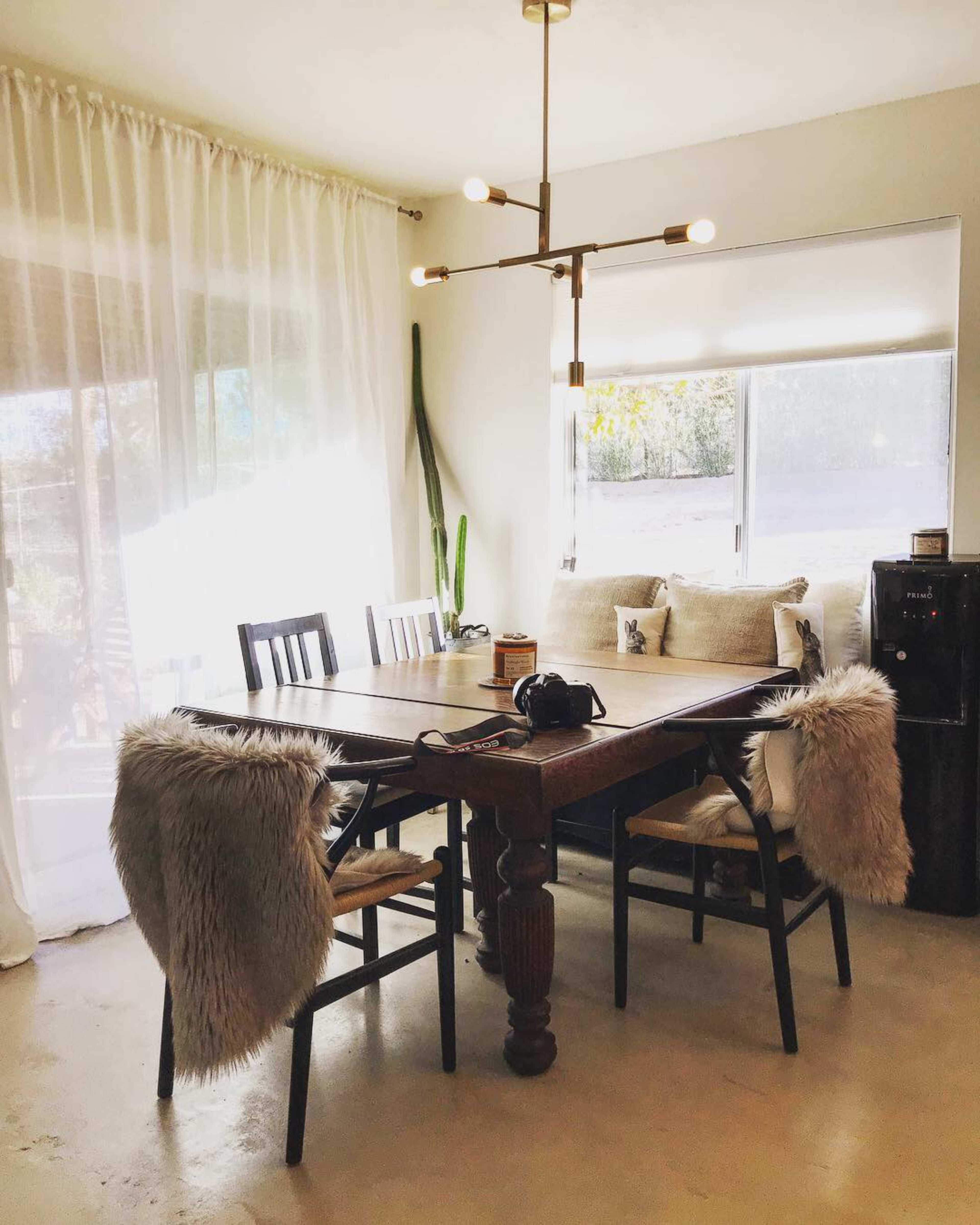 A wooden dining table with four chairs, two of which have fur throws, is set near a large window with sheer curtains and a cushioned bench.