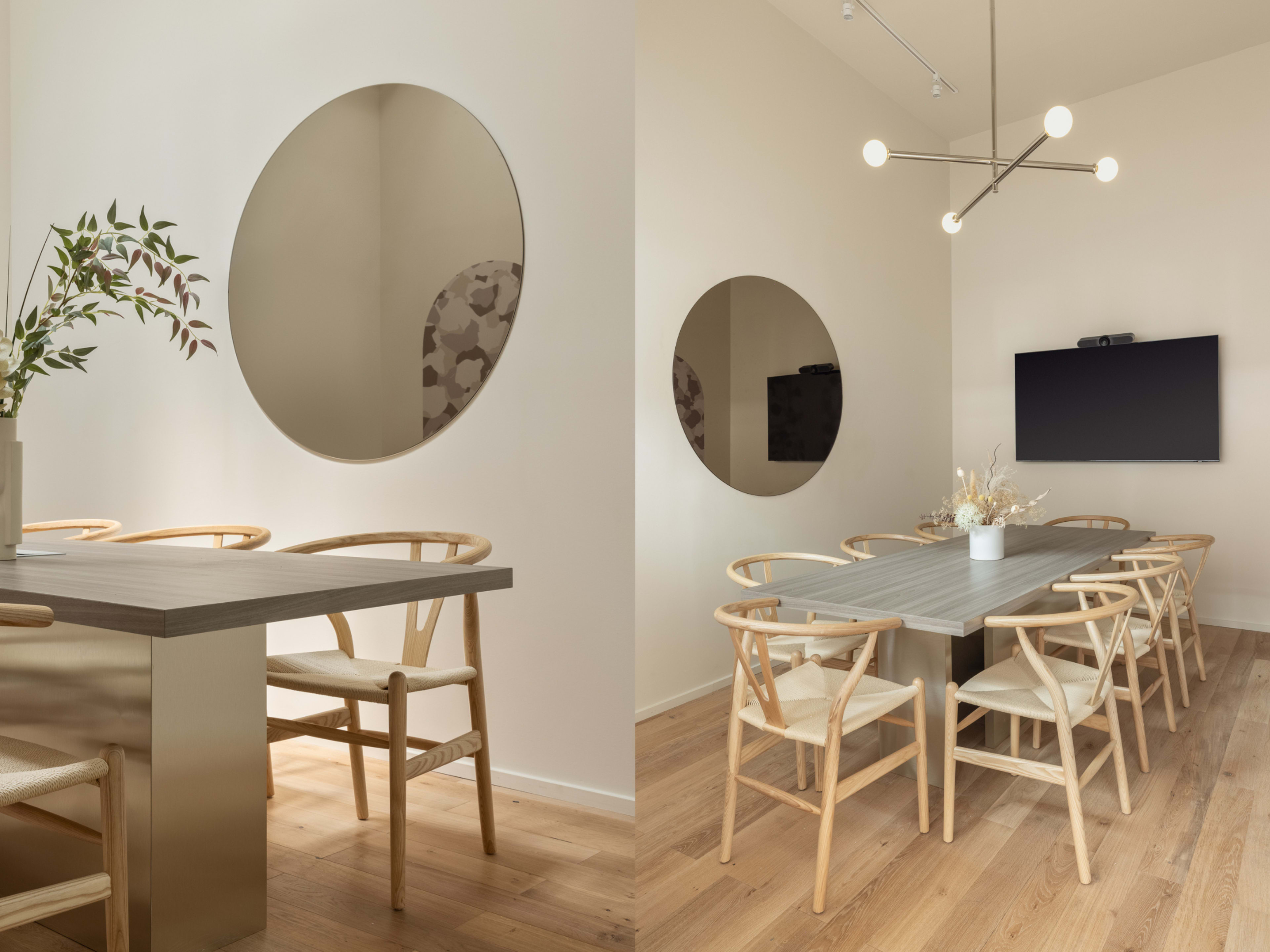 The image features a modern dining area with a large oval mirror, a simple wooden table, and several light-colored chairs arranged around it, alongside a wall-mounted television.