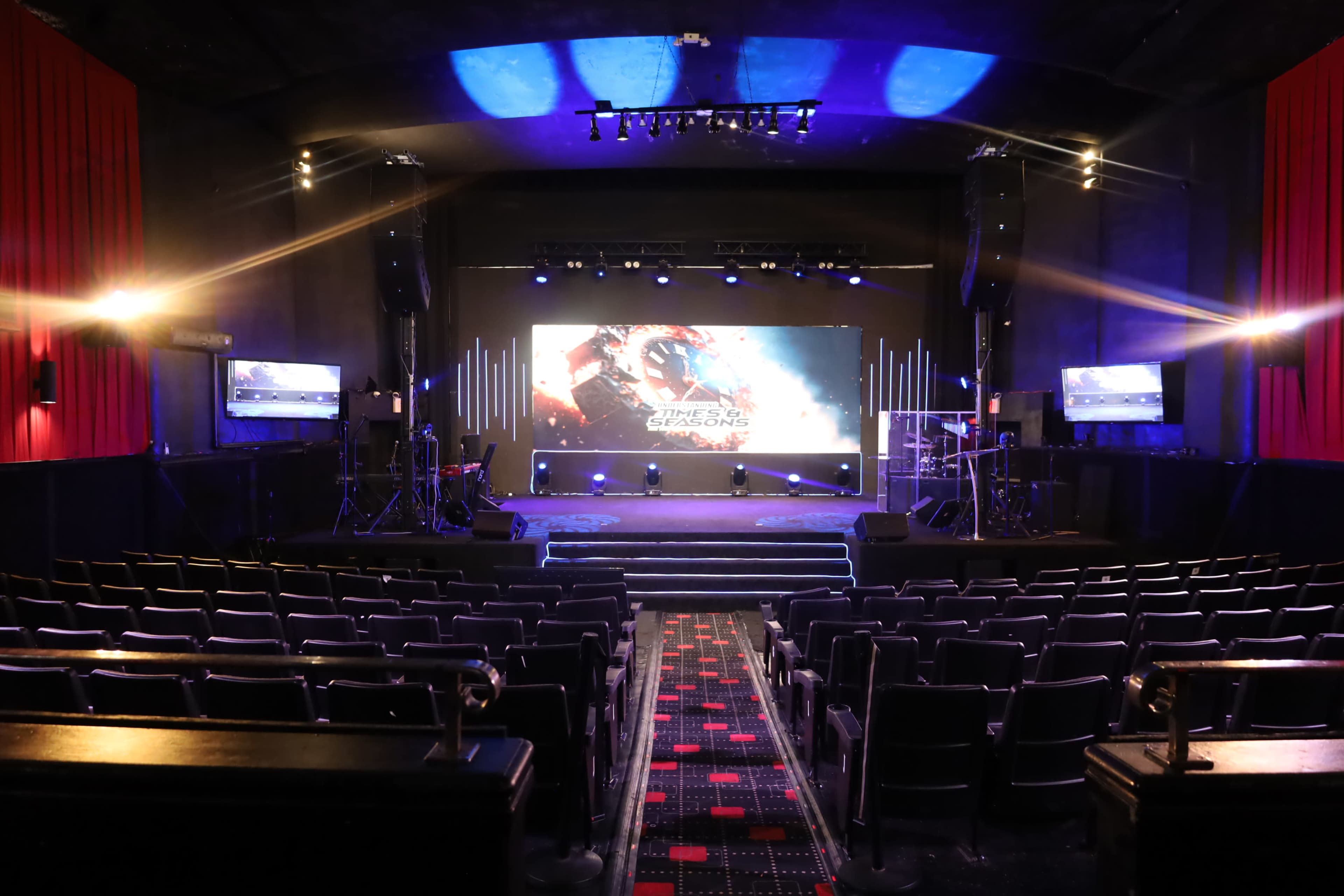 The image shows a stage in a theater with a large screen at the back and rows of empty seats in front.