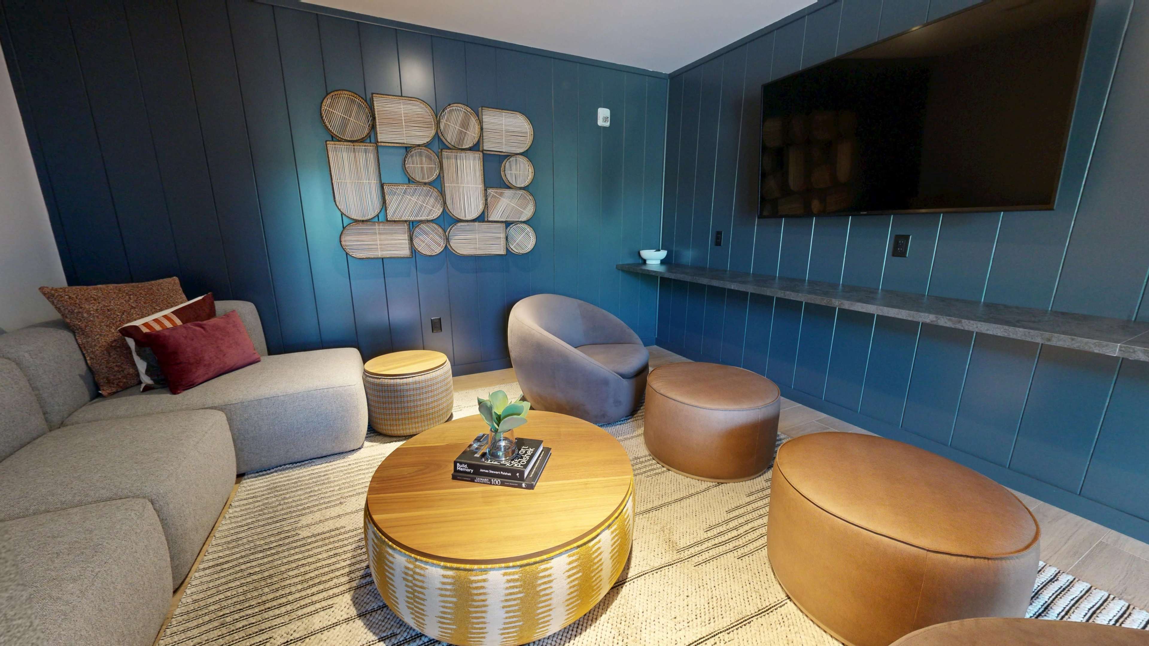 The image shows a modern, styled living area featuring a gray sectional sofa, brown ottomans, a round wooden coffee table, and a wall adorned with decorative wall art and a mounted television.