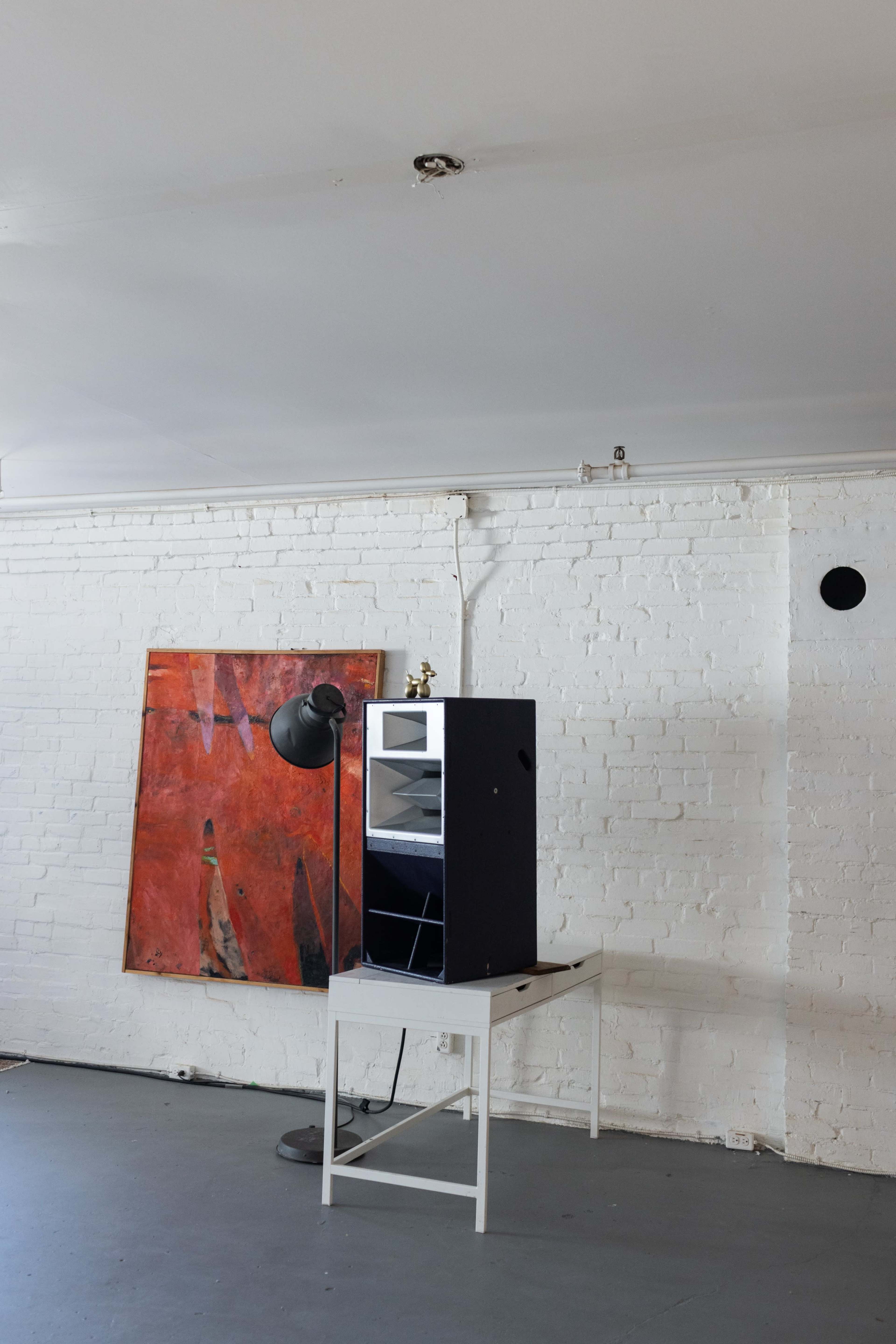 The image shows an empty, minimalist room with a white brick wall, a large abstract painting, a small glass cabinet on a table, and a floor lamp.