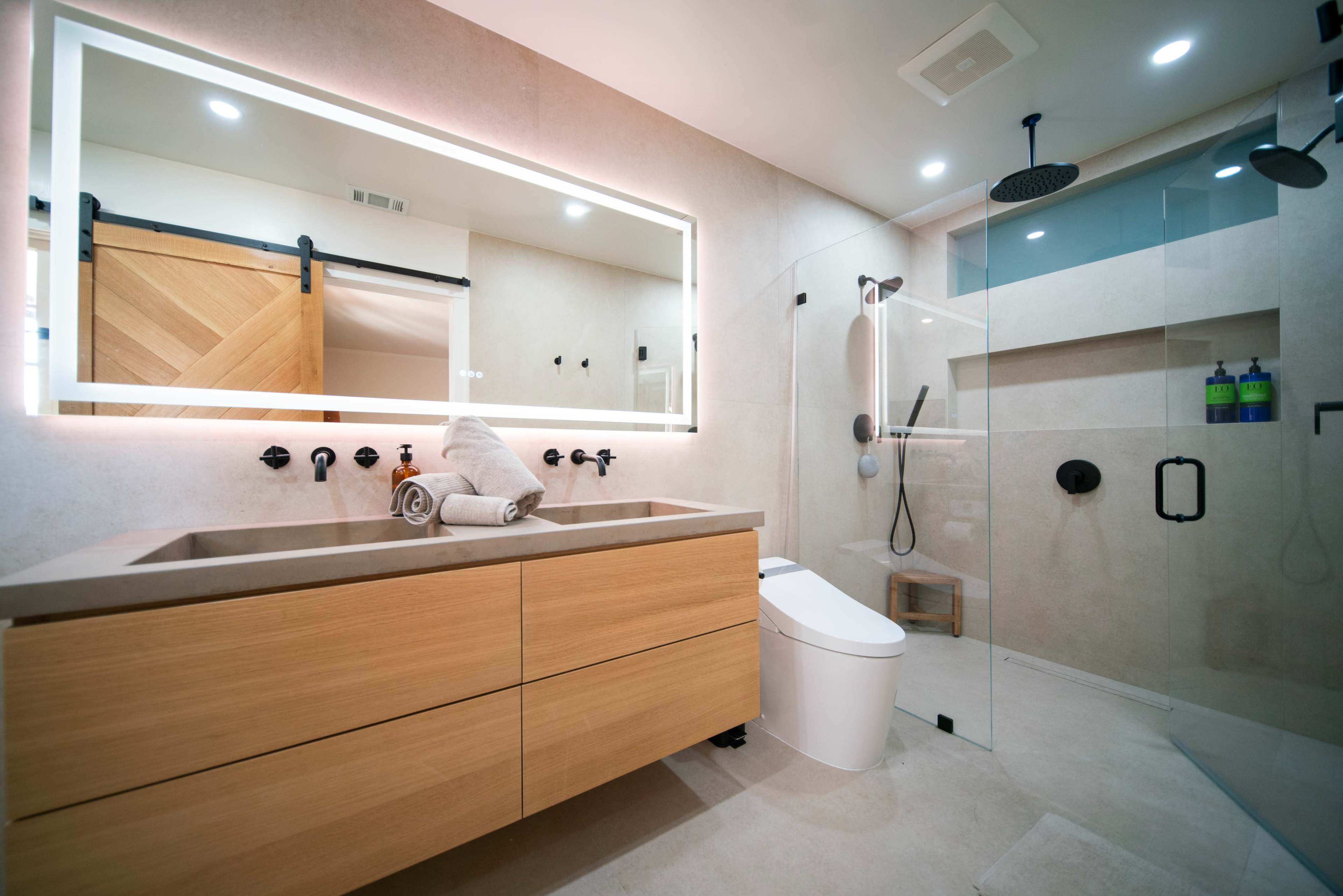 The image shows a modern bathroom featuring a glass shower, a wooden vanity with a sink, and a backlit mirror.