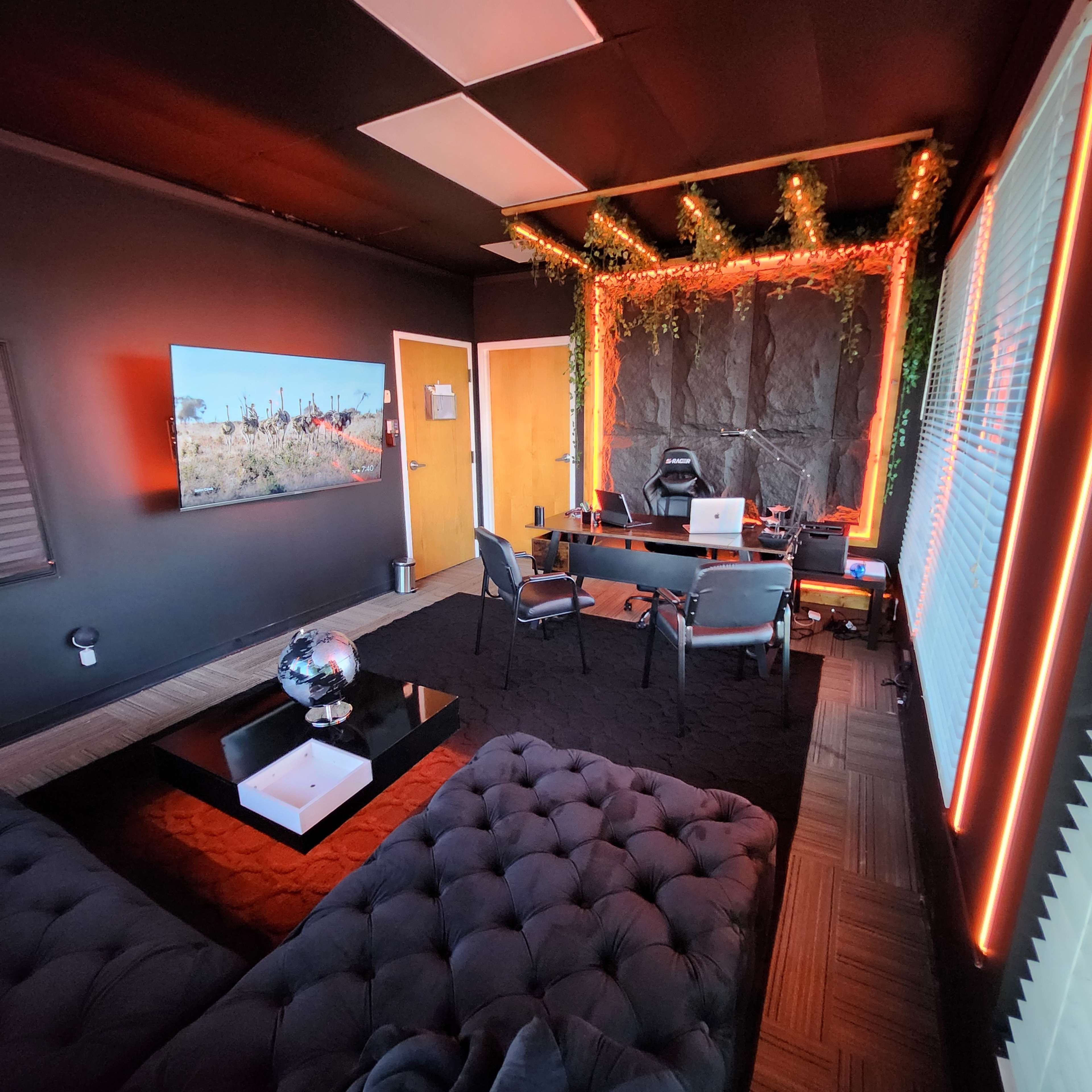 The image shows a modern office space with dark walls, a large screen displaying an outdoor scene, a black sofa, and a desk with a laptop, surrounded by ambient lighting and decorative plants.