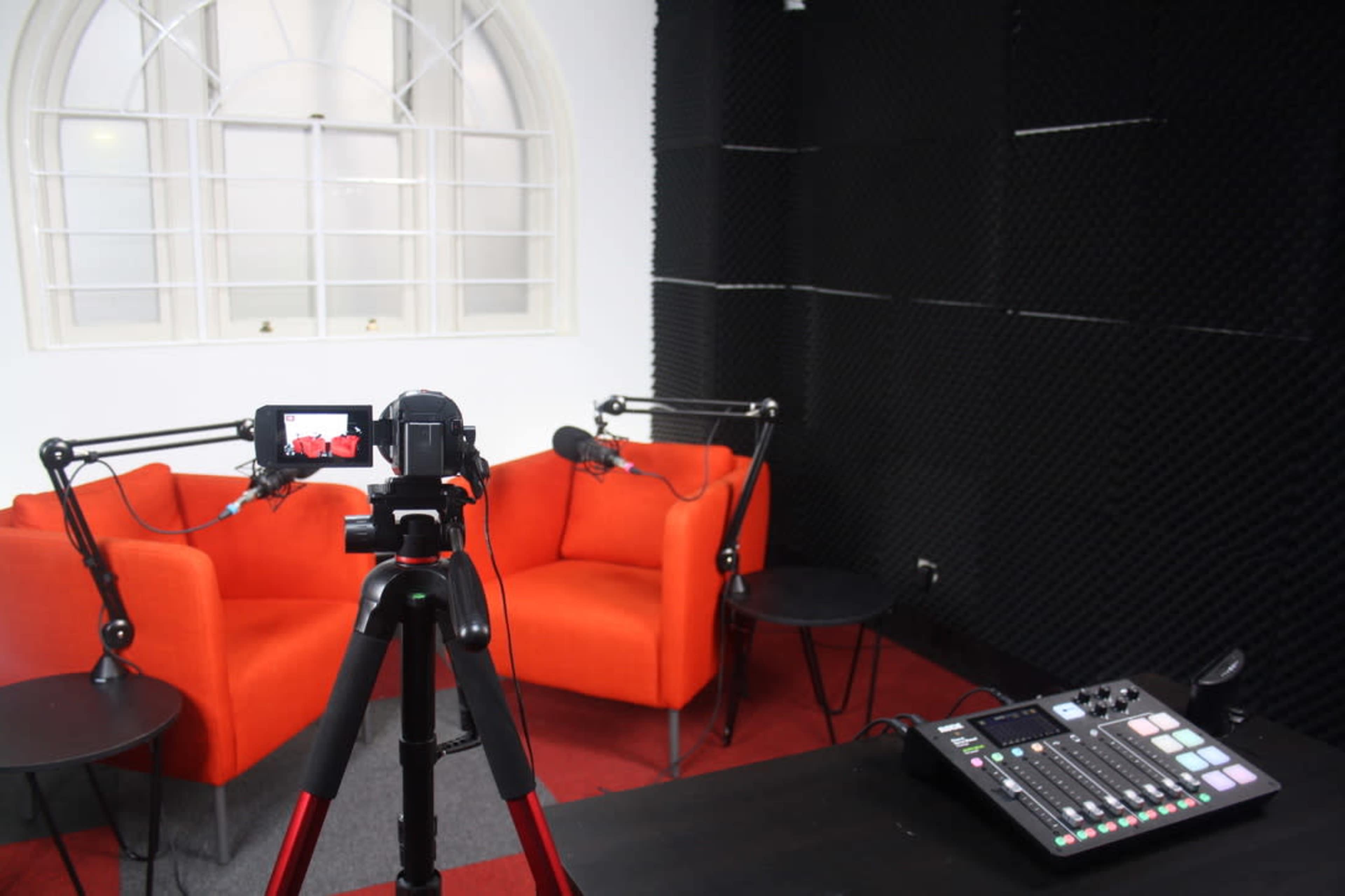 A recording studio with two orange chairs, a microphone setup, and a video camera on a tripod in front of a control panel.