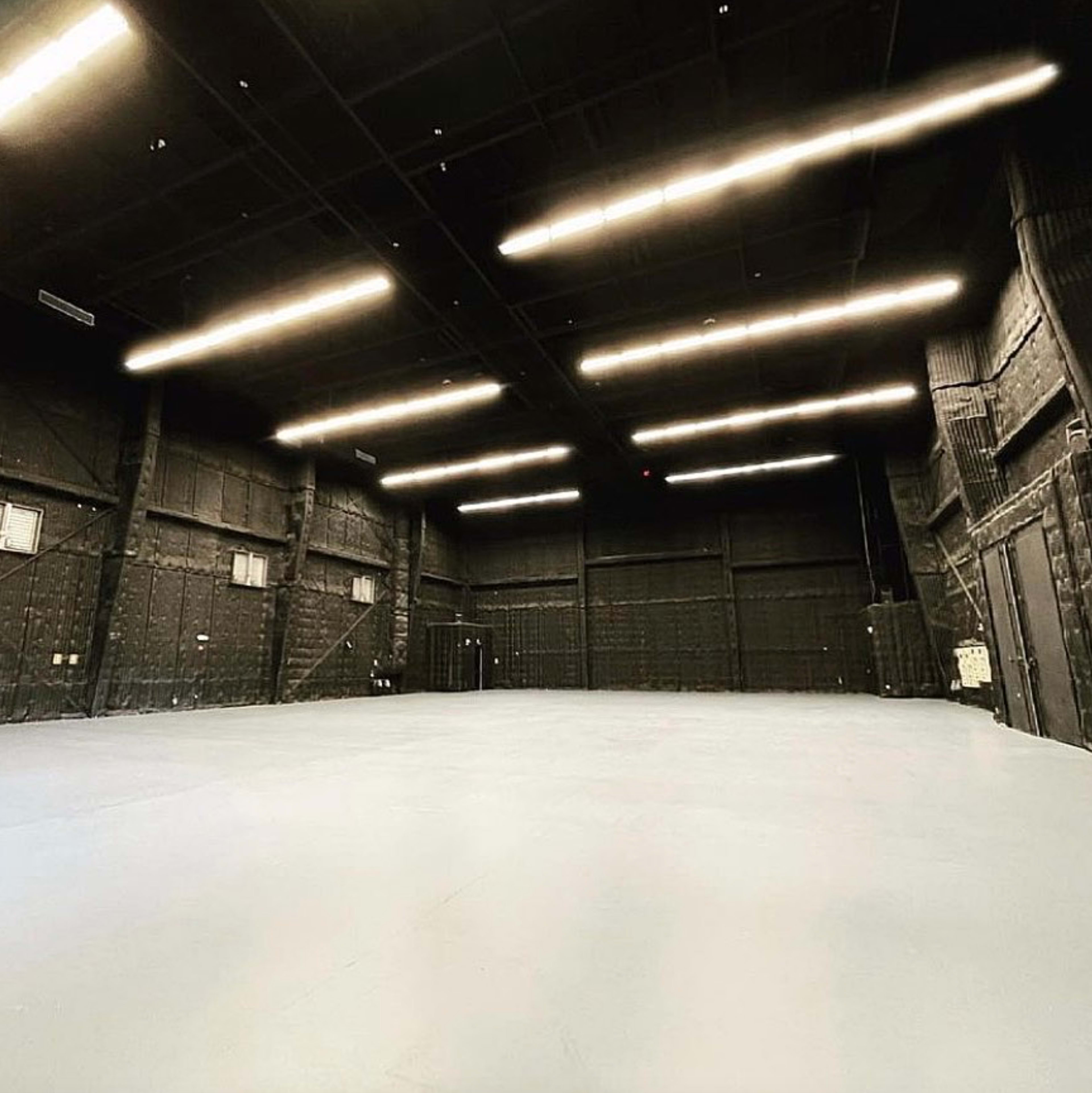 A large, empty warehouse space features dark walls and a smooth, light-colored floor, illuminated by overhead strip lights.