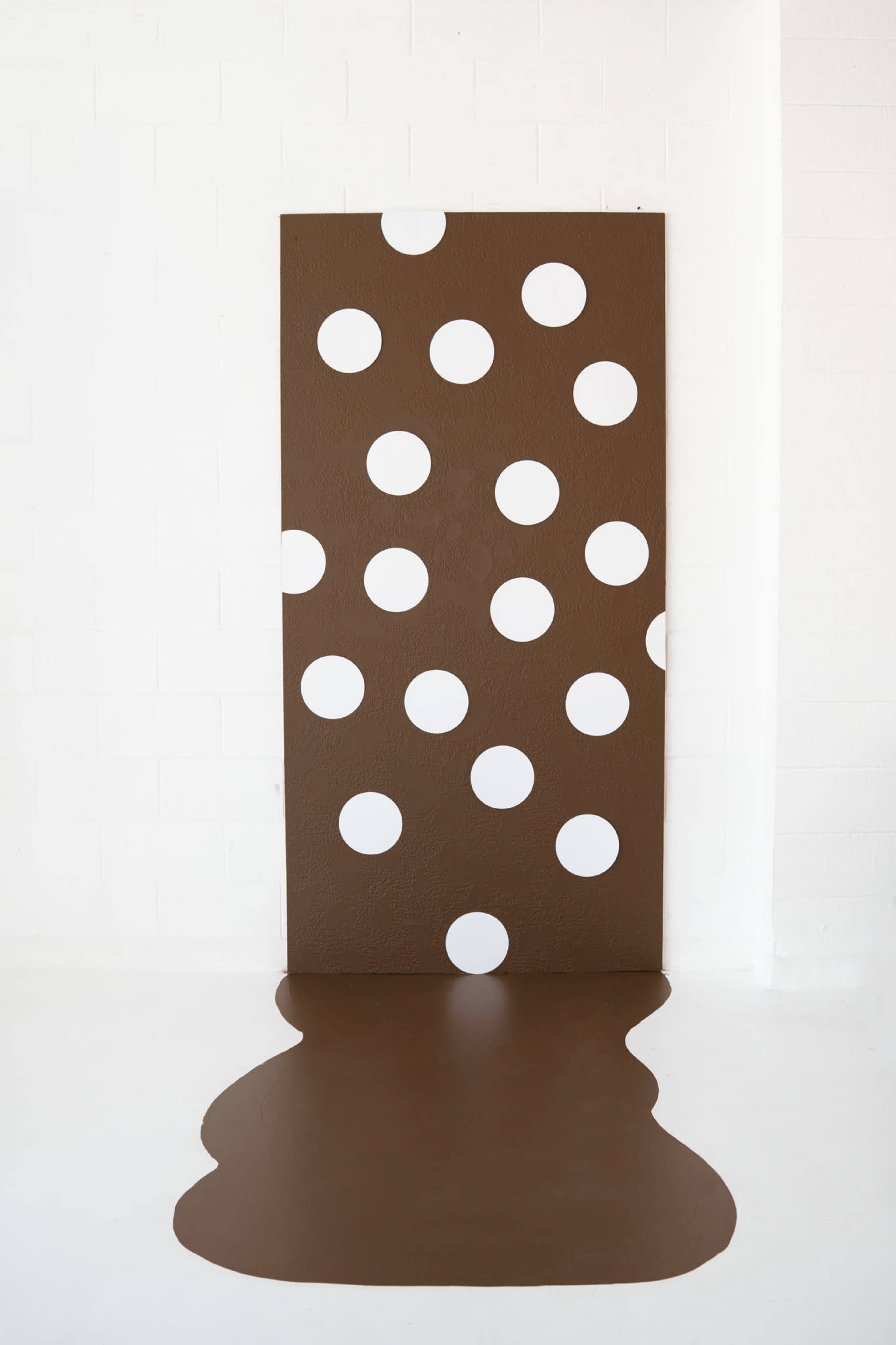 The image features a large brown panel with white polka dots, positioned against a white wall, with a brown floor section appearing to drip from the base of the panel.