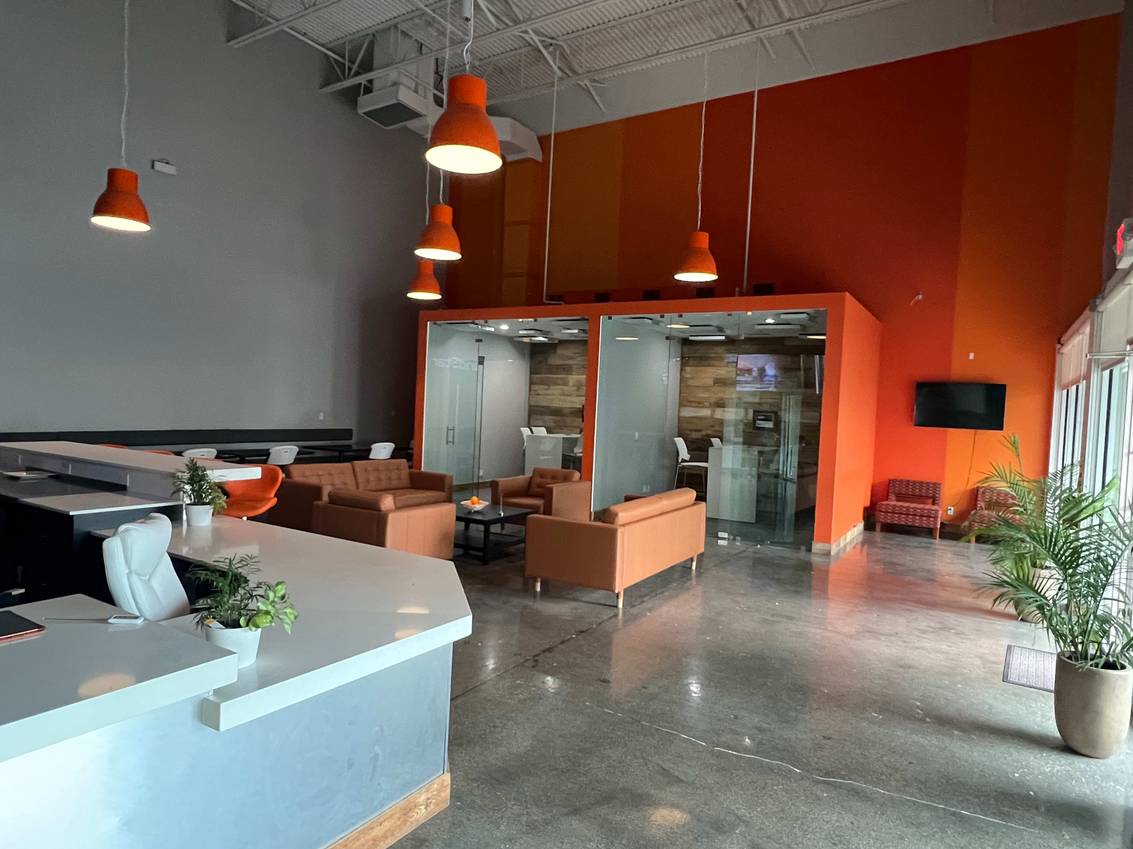 The image shows a modern reception area with a front desk, orange pendant lights, and a seating area featuring brown sofas and glass-walled conference rooms.