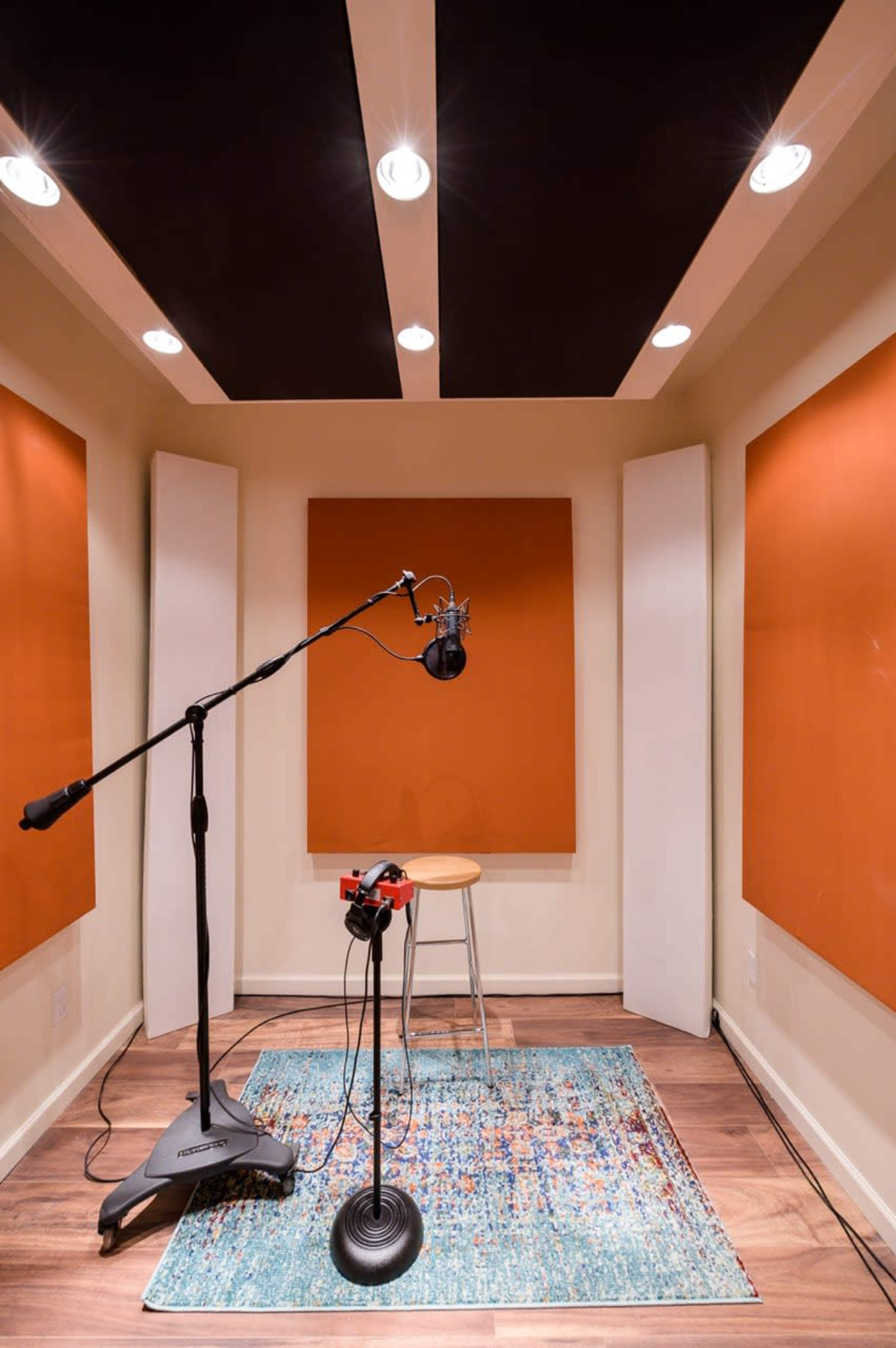 The image shows a small recording studio with orange soundproofing panels, a microphone on a stand, a stool, and an area rug on the wooden floor.