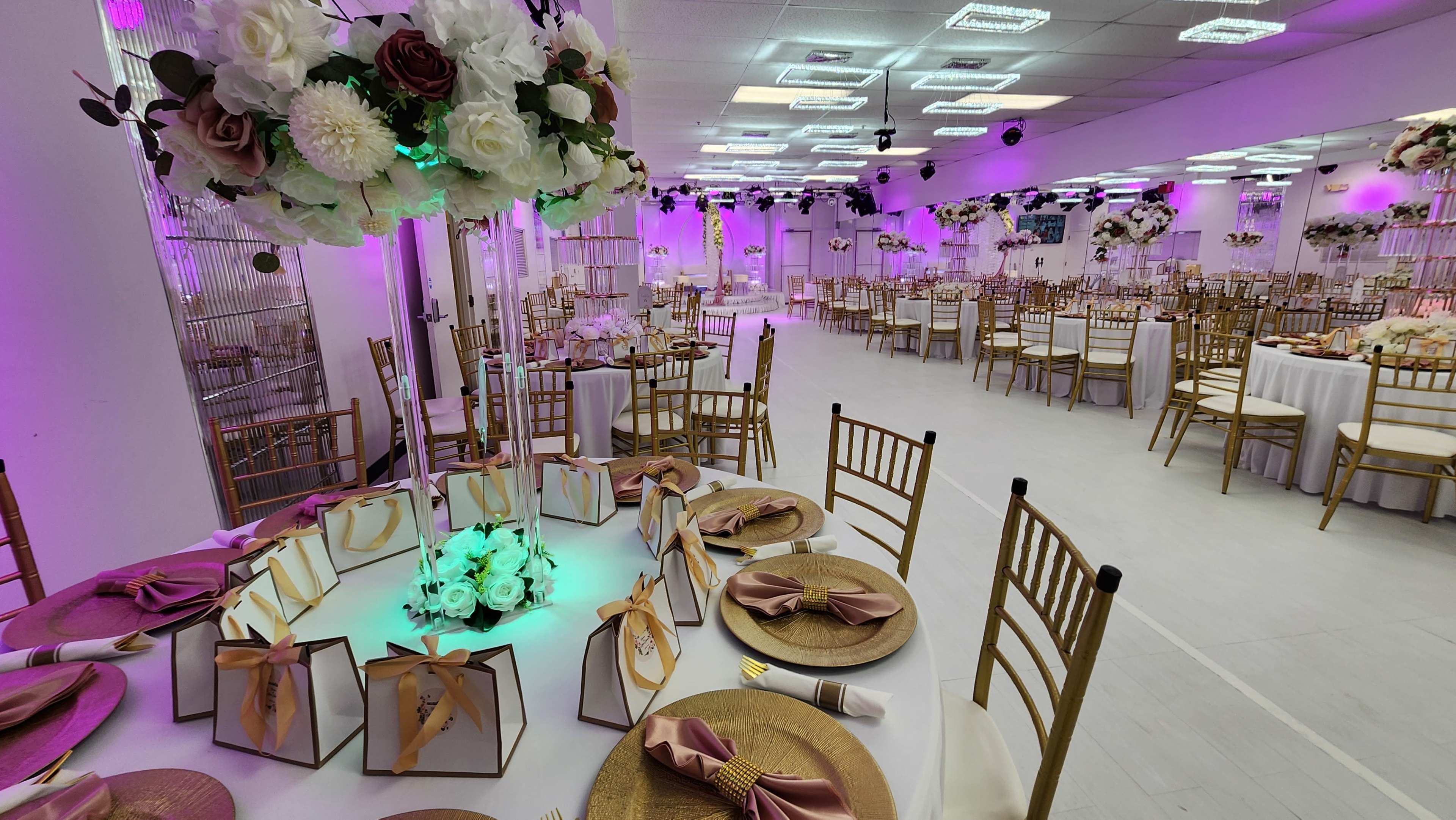 A banquet hall is set up for an event, featuring elegantly arranged tables with gold chairs, floral centerpieces, and ambient purple lighting.