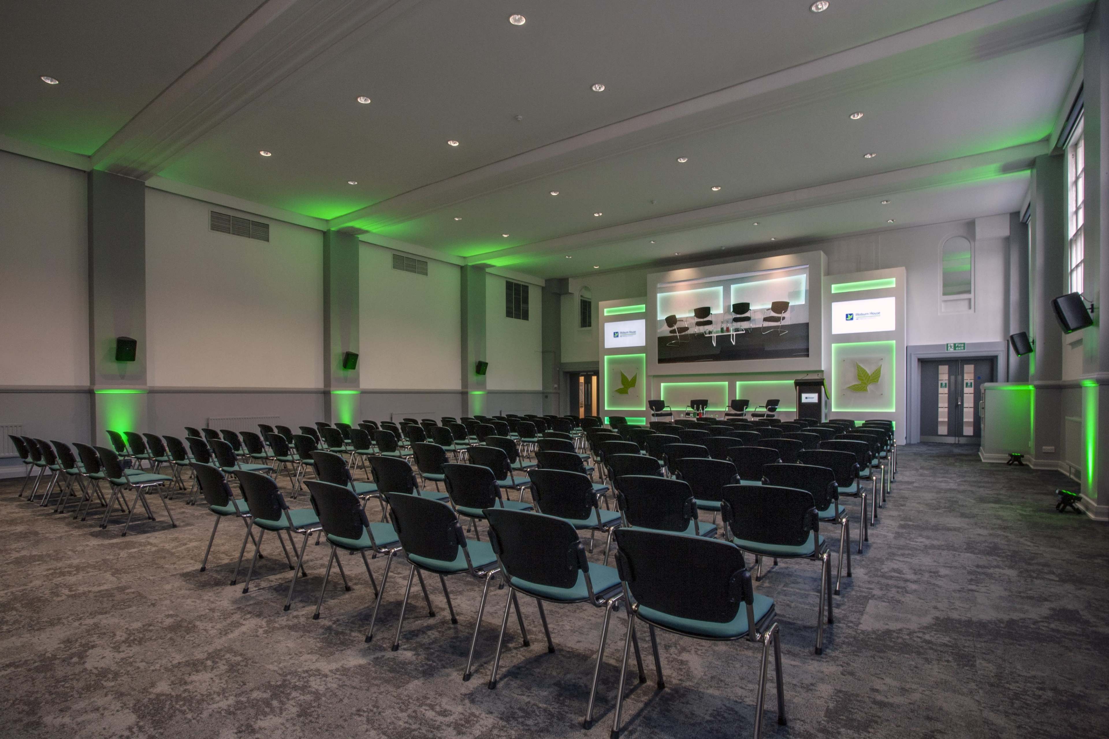 A large conference room with rows of black chairs facing a central stage that features a projection screen and multiple displays.