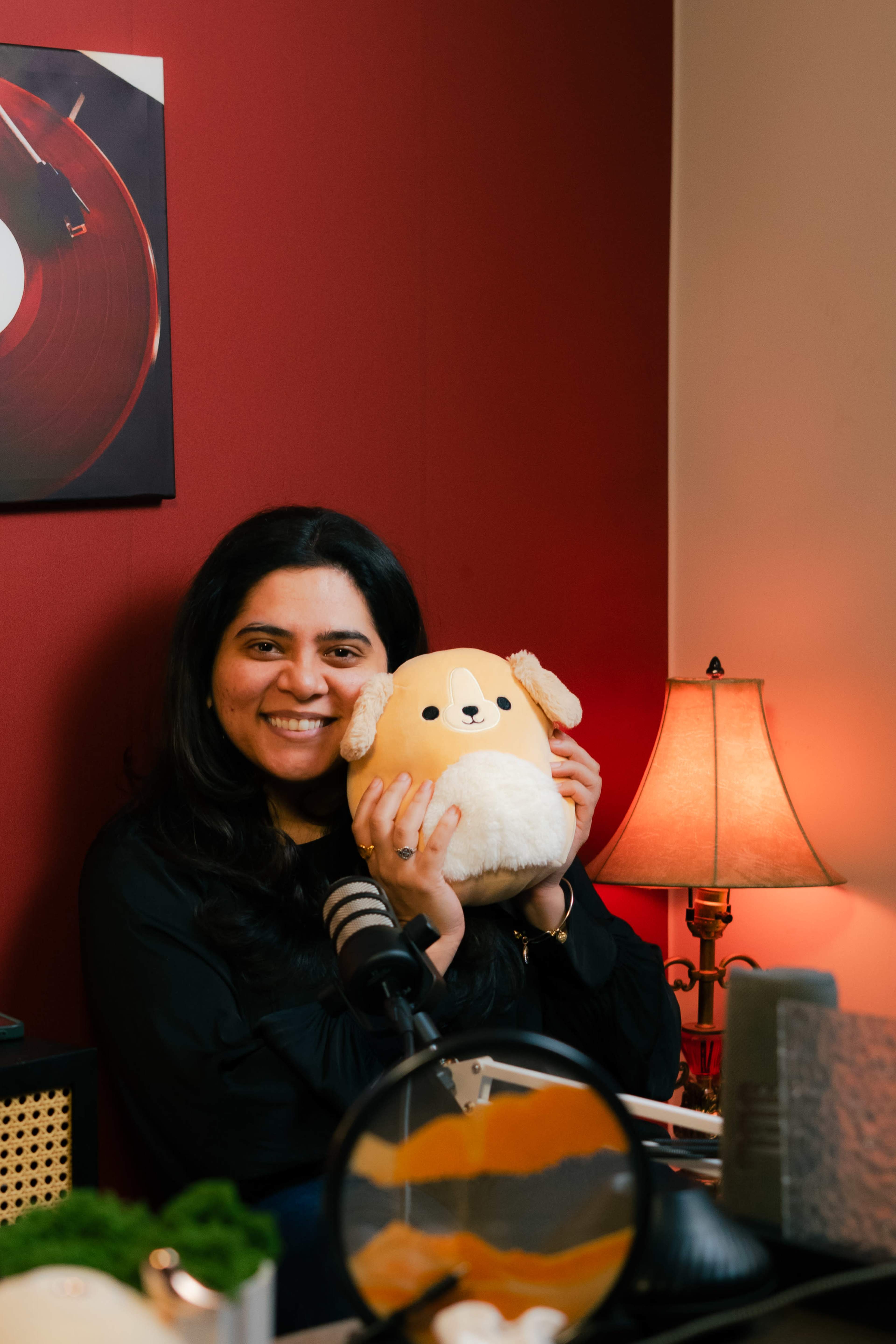 A woman holds a plush dog toy while sitting at a desk with a lamp and various items in a room with red walls.