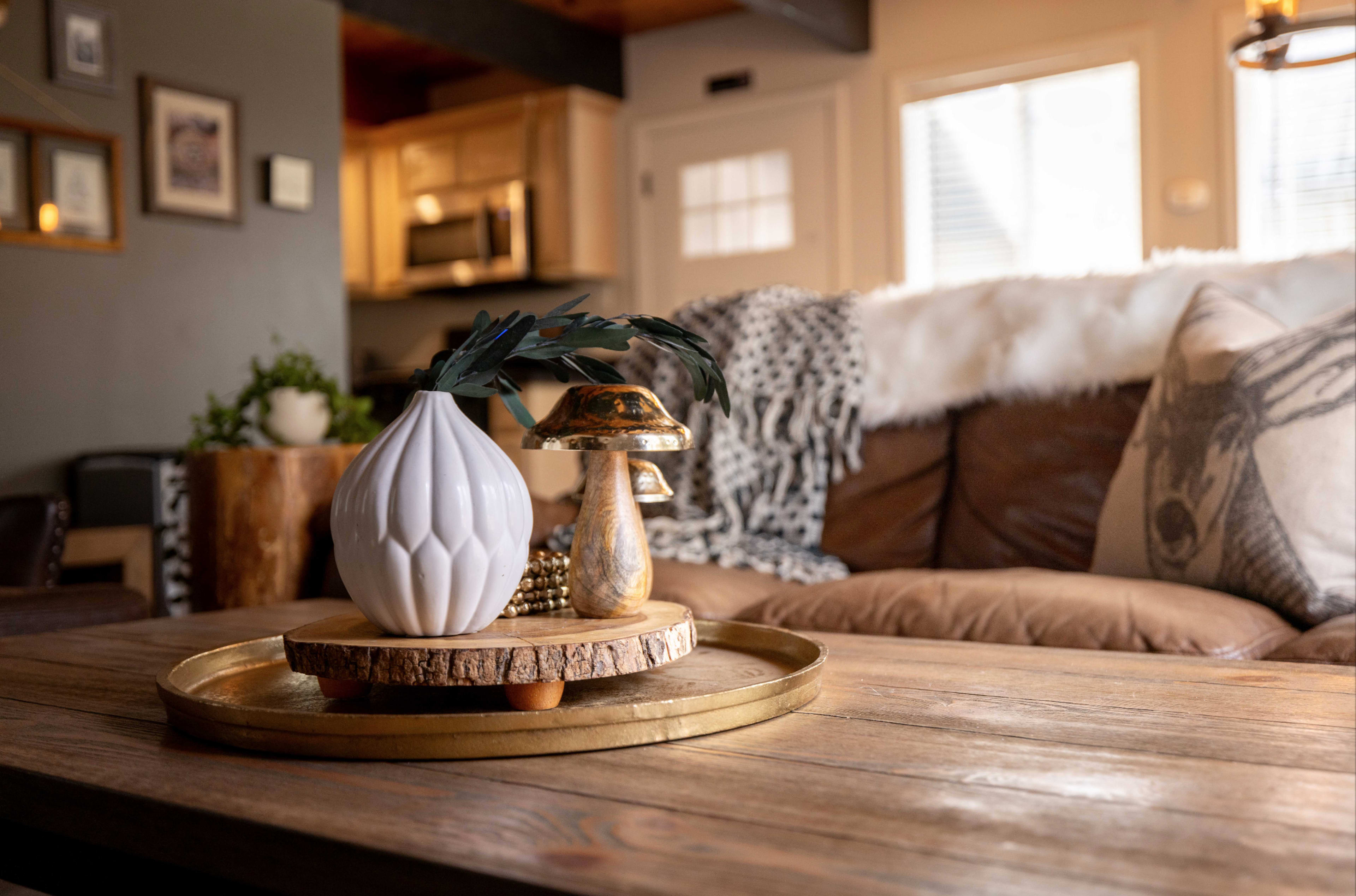 A coffee table features a decorative white vase and a golden mushroom sculpture, set against a backdrop of a cozy living room with a leather sofa and window light.