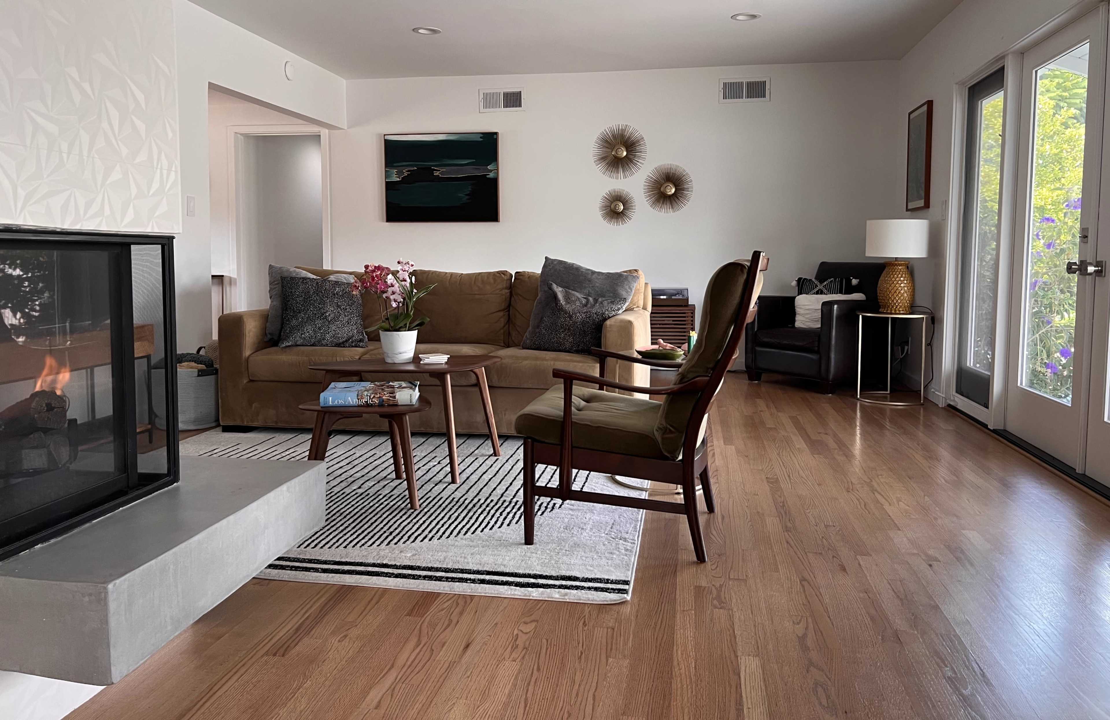 A modern living room featuring a brown couch, a wooden chair, a coffee table with books, and large windows that allow natural light to enter.