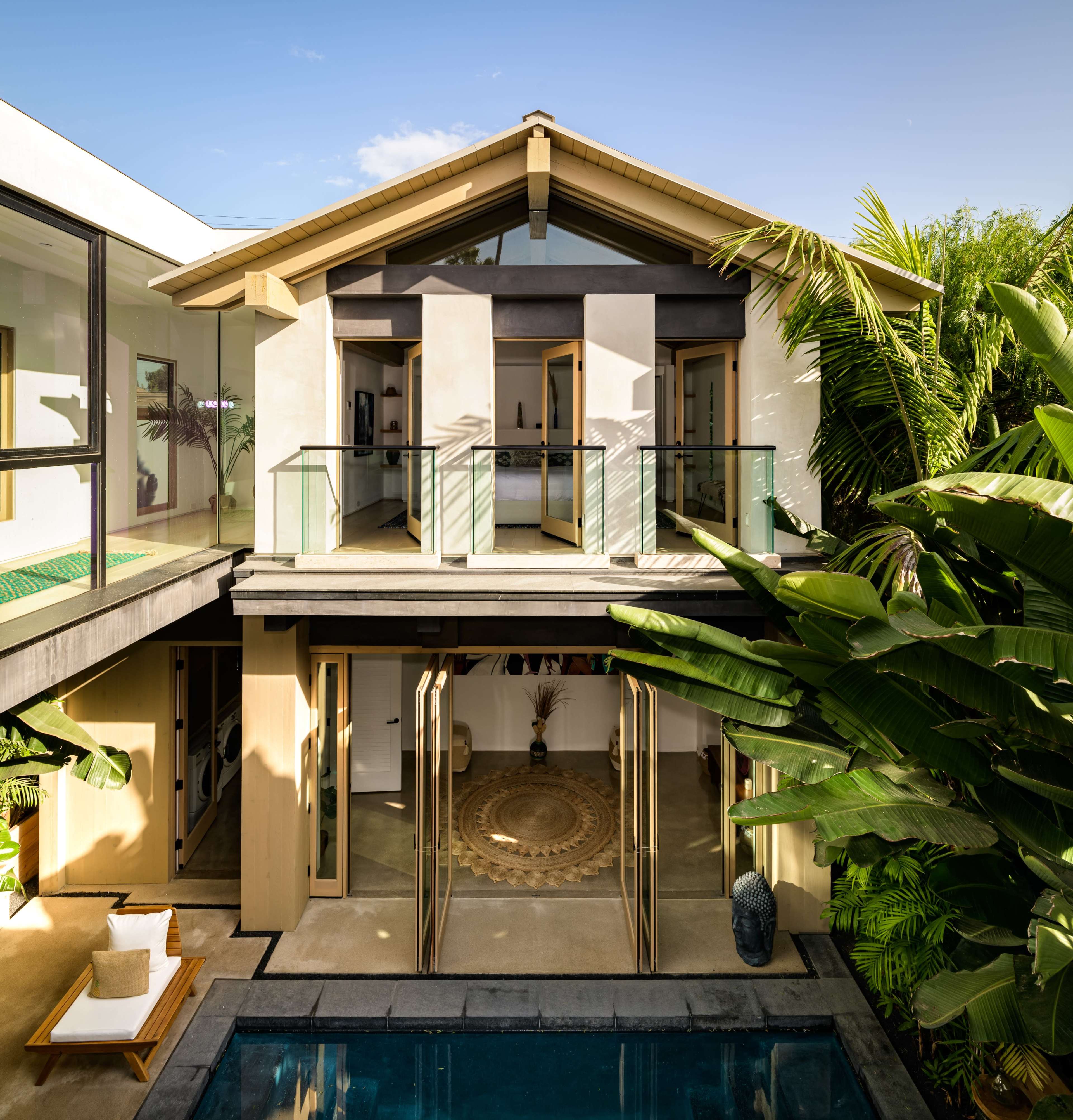 A modern house with a sloped roof features large glass doors and windows, surrounded by lush tropical plants and a swimming pool.