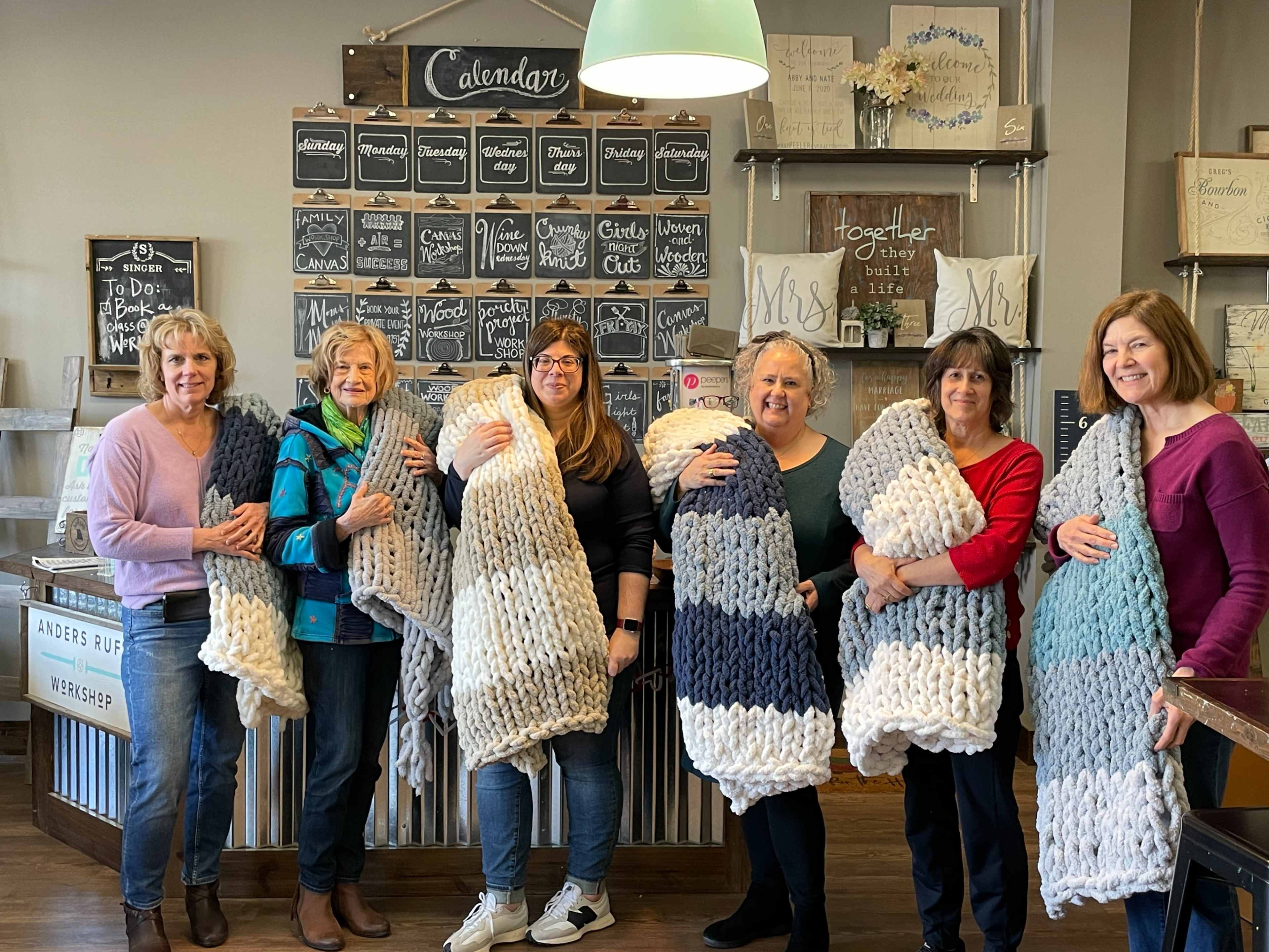 A group of six women is standing in a craft store, each holding a large, knitted blanket.