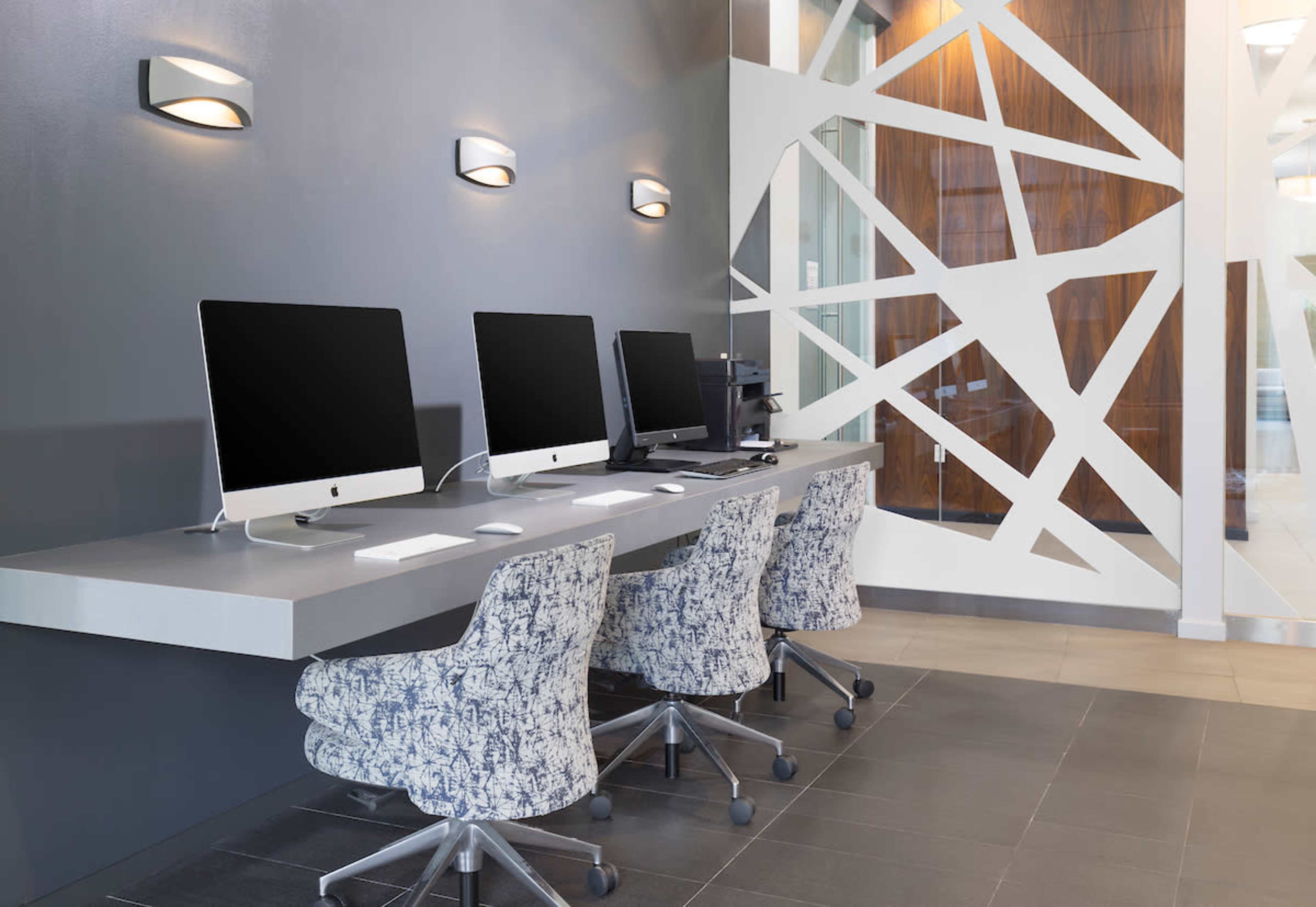 Three desktop computers are positioned on a sleek, light-colored desk, accompanied by patterned chairs, against a modern gray wall.