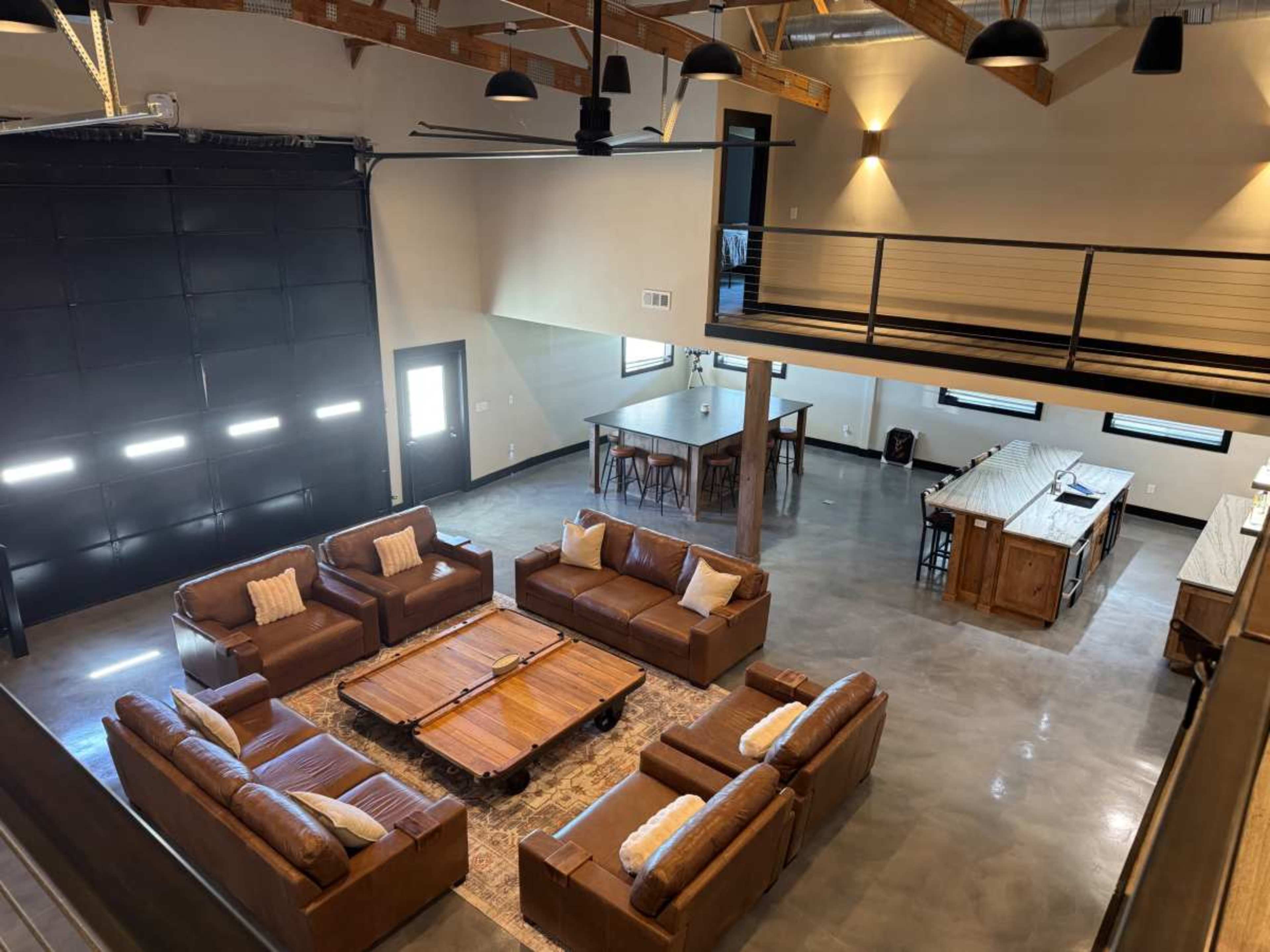 The image shows a spacious, modern living area with a large sectional sofa, a wooden coffee table, and a dining area featuring a high table and chairs, all under a high ceiling with exposed beams.