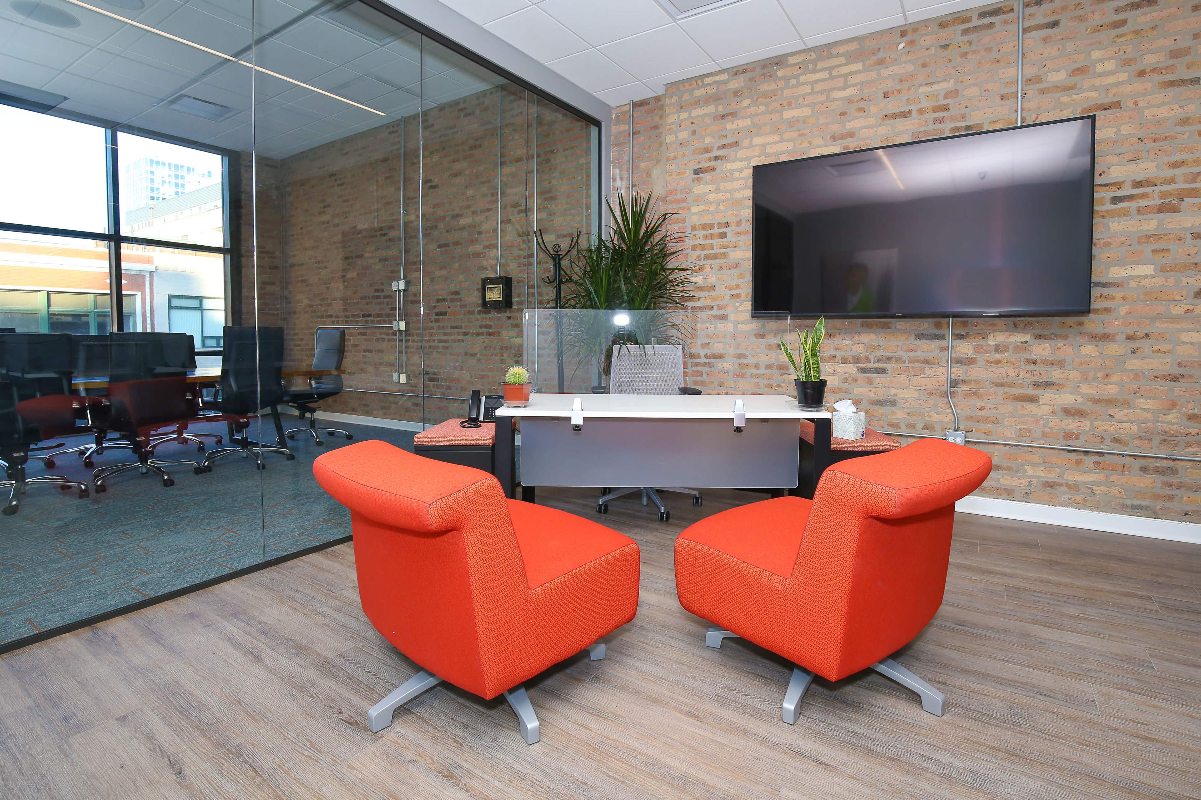 A modern office space with two orange chairs facing a sleek desk, a large television mounted on the wall, and a glass partition revealing a conference room.