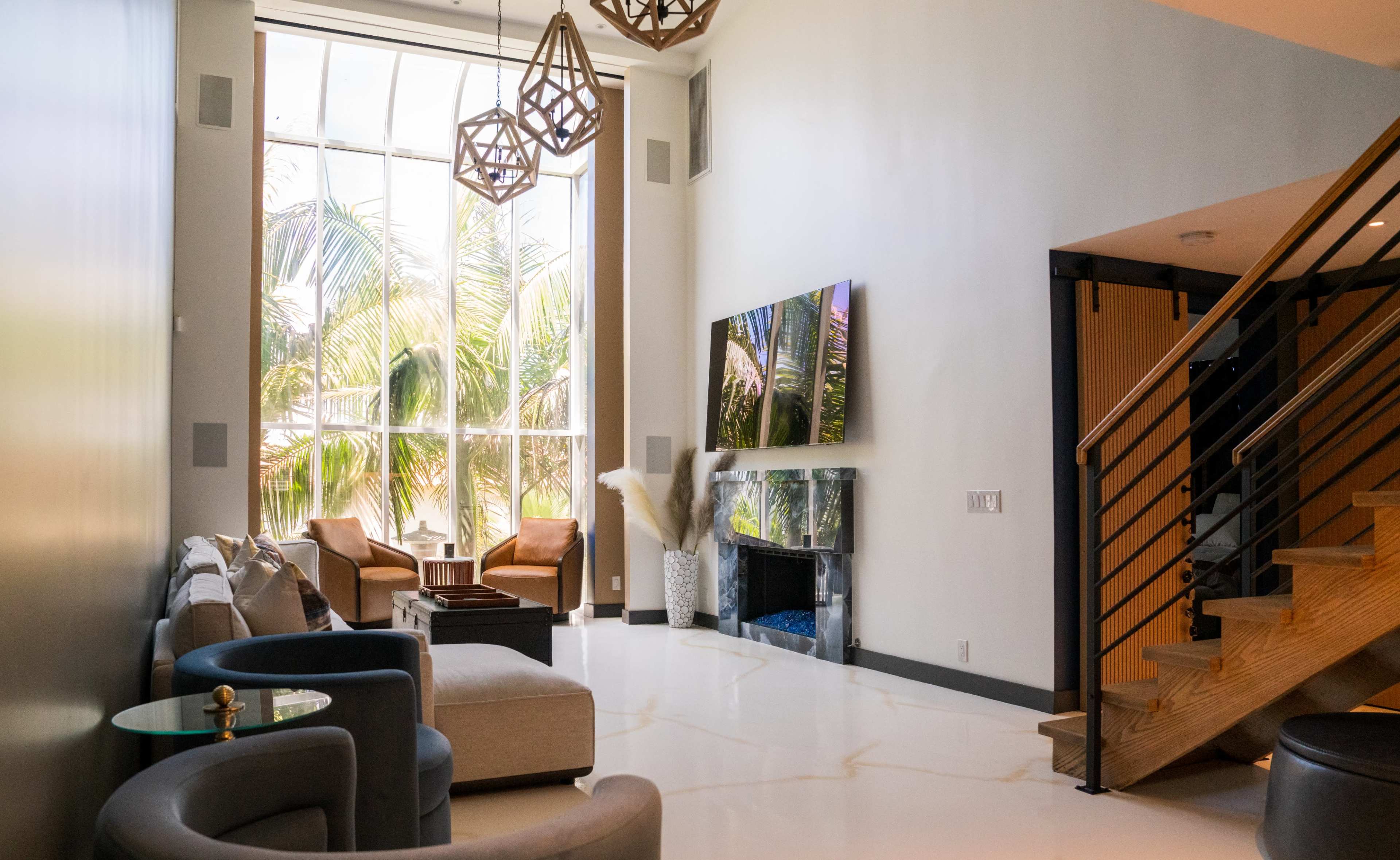 The image shows a modern living room with large windows, a flat-screen TV on the wall, and a staircase leading to an upper level.