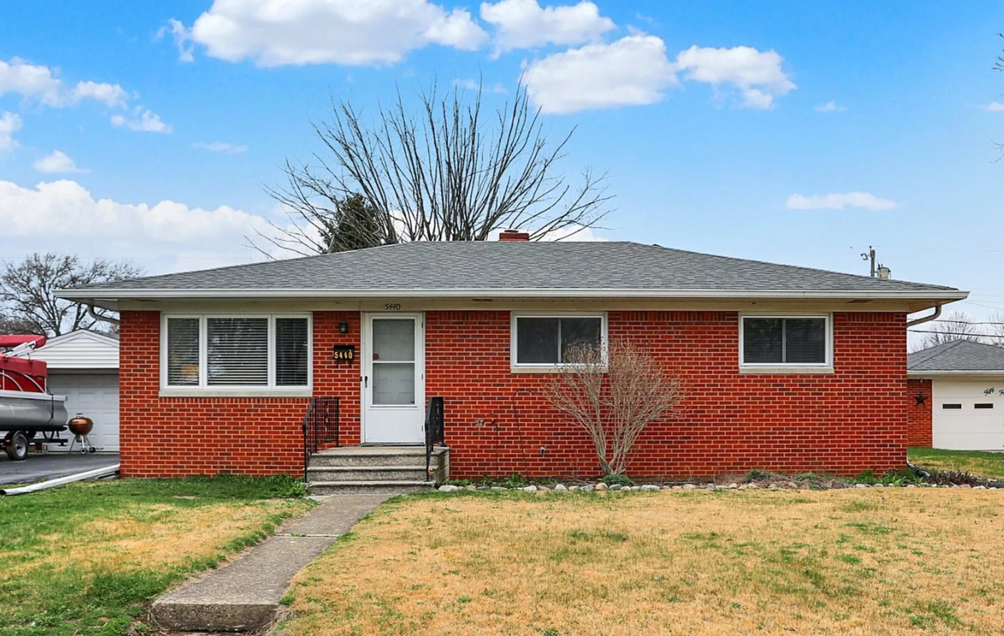 The image shows a single-story, brick house with a simple design, featuring a concrete walkway leading to the front door and a sparse lawn.