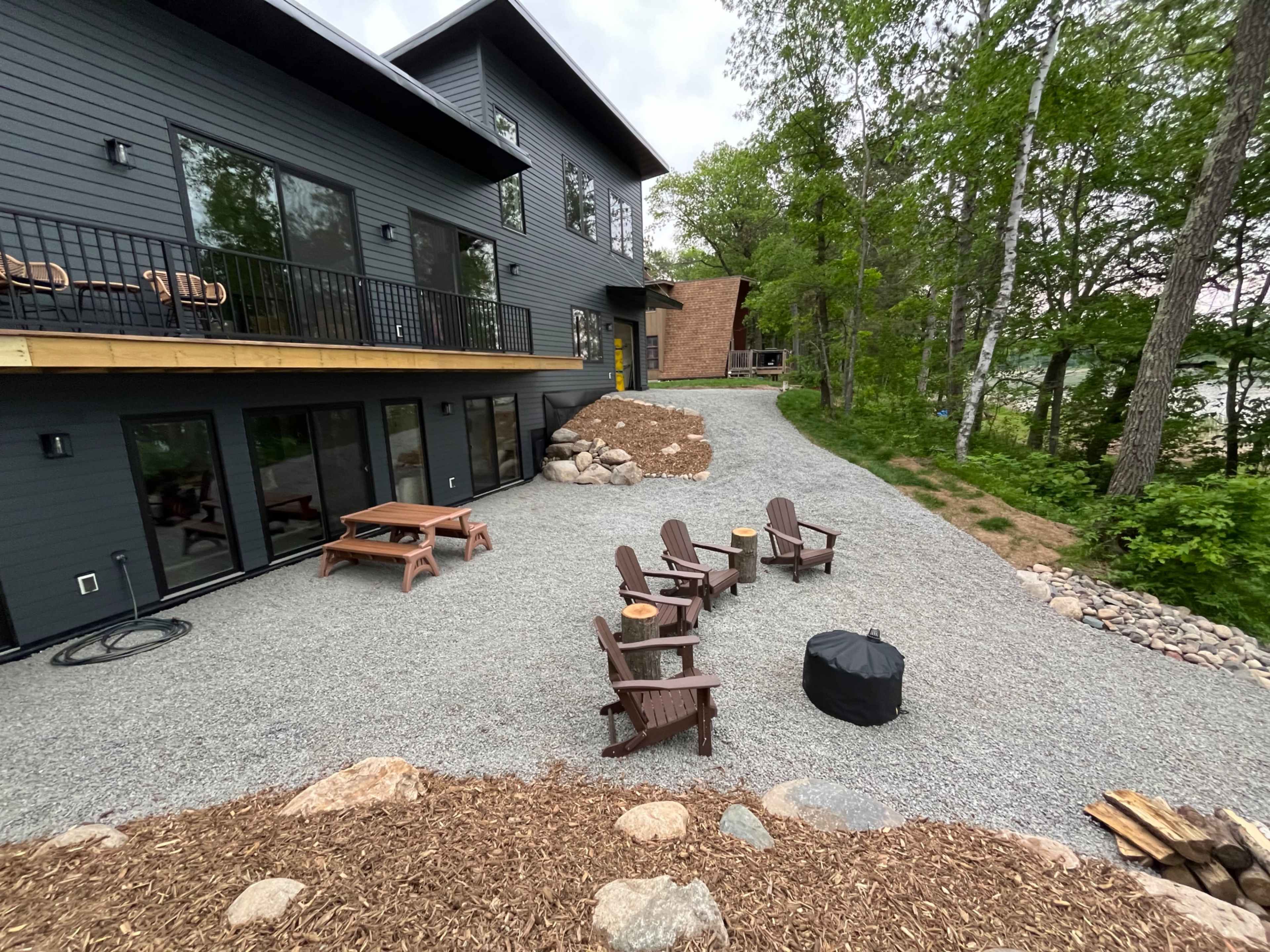 The image shows a gravel patio with several wooden chairs and a fire pit in front of a modern two-story house surrounded by trees.