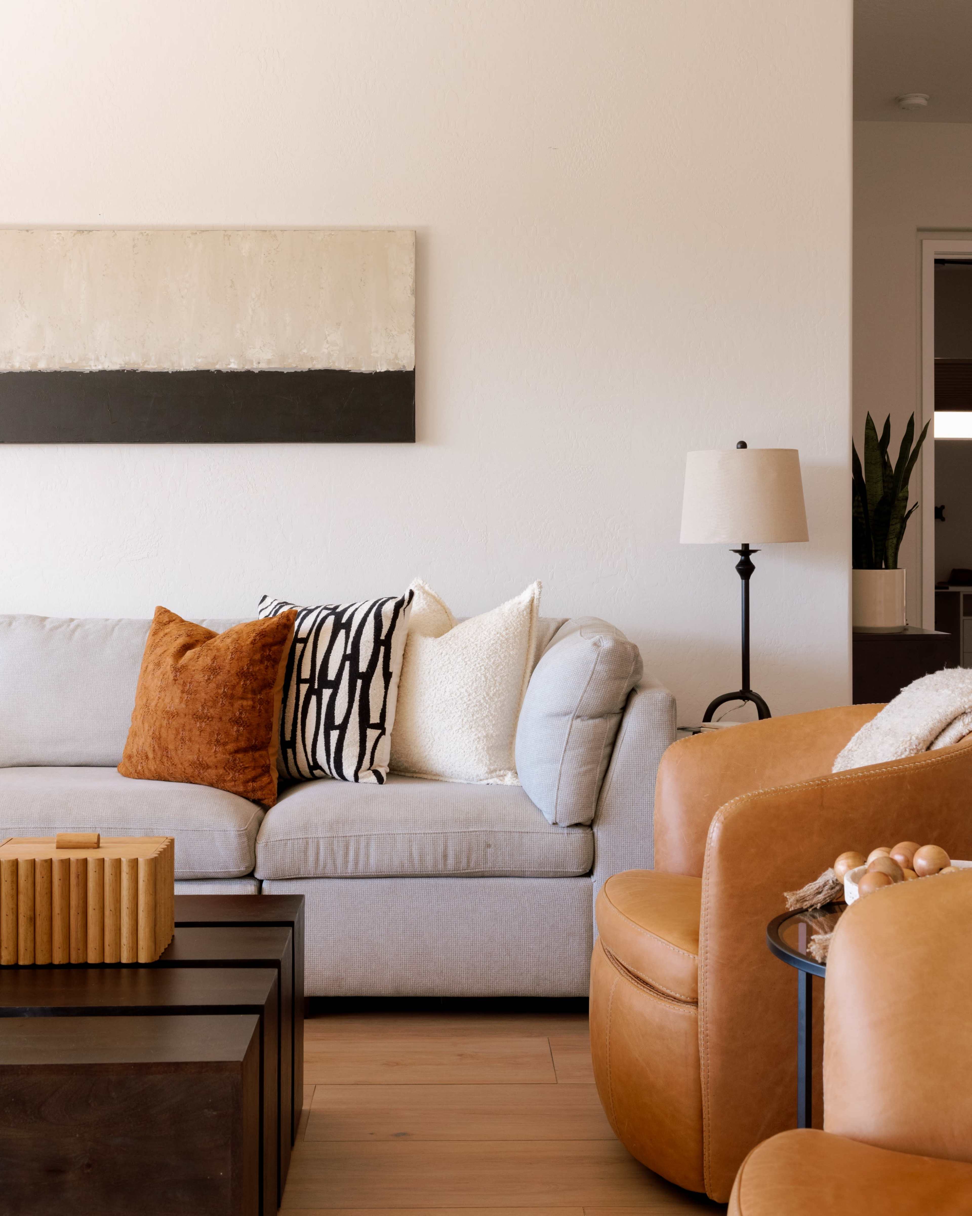 The image shows a modern living room featuring a light gray sofa with decorative pillows, a brown leather armchair, a wooden coffee table, and a lamp beside the sofa.